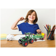Smiling child in blue shirt with Alexander Tractor Construction Set on wooden desk, colorful pencils in holder nearby.