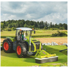 Claas toy tractor with front disc mower on green field, featuring realistic details and vibrant colors, against a forest backdrop.