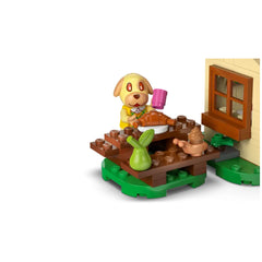 LEGO minifigure of Goldie, the dog, sitting at a picnic table with a croissant, pear, and drink near her cozy house.