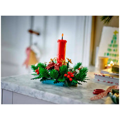 A vibrant LEGO Christmas table decoration featuring a tall red candle surrounded by lush green foliage, golden stars, and red berries, set on a marble surface.