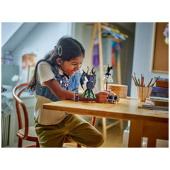 A child smiles while building LEGO® figures of Maleficent and Cruella De Vil on a wooden table, surrounded by art supplies.
