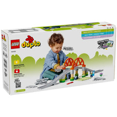 A toddler in a blue shirt and denim jacket plays with the LEGO® DUPLO® Train Bridge and Tracks Expansion Set, featuring a raised bridge, track pieces, and colorful blocks. The child engages creatively, enhancing motor skills and imagination in a vibrant play scene.