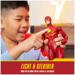 A young boy in a blue top points at a red Flash action figure with lights and sounds, showcasing excitement in a cozy indoor
