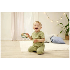 Eichhorn Baby HiPP Sound Rattle is held by a smiling child in a green outfit, sitting on a white rug in a bright room.