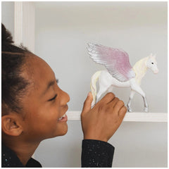 A young girl smiles while holding a white and pink winged horse figurine indoors against a white wall.
