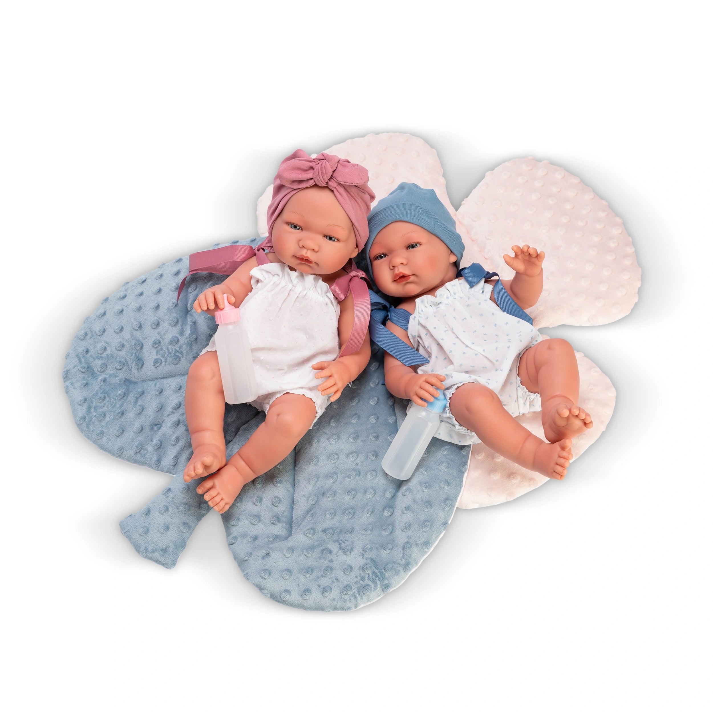 Two ASI Maria dolls in rompers with matching hats, holding bottles, lying on a soft, floral textured pillow.