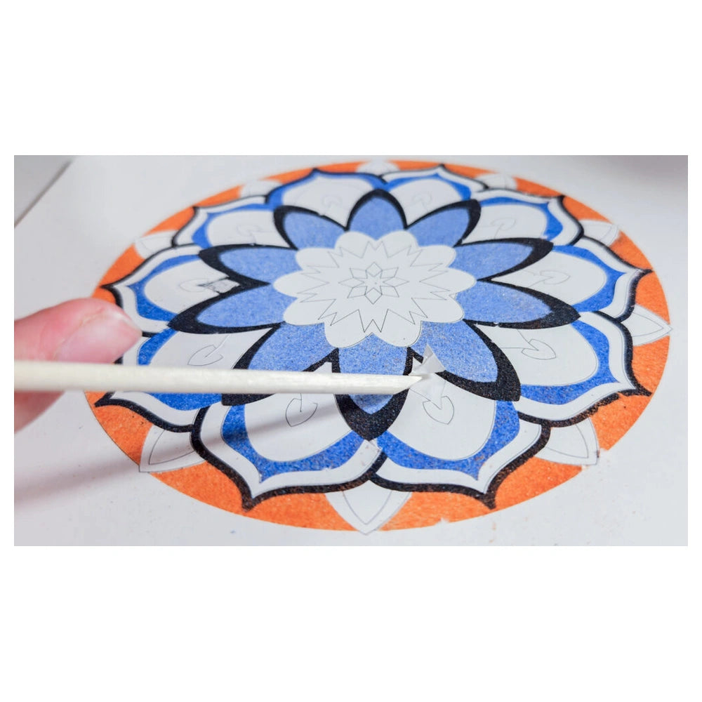 Sand art mandala features a colorful pattern with blue and orange concentric circles, as a hand holds a paintbrush above it.