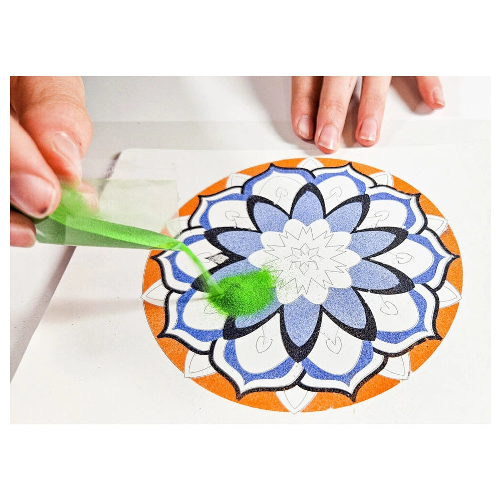 Sand art mandala craft in progress with hands applying green substance to a symmetrical blue and white design on paper.
