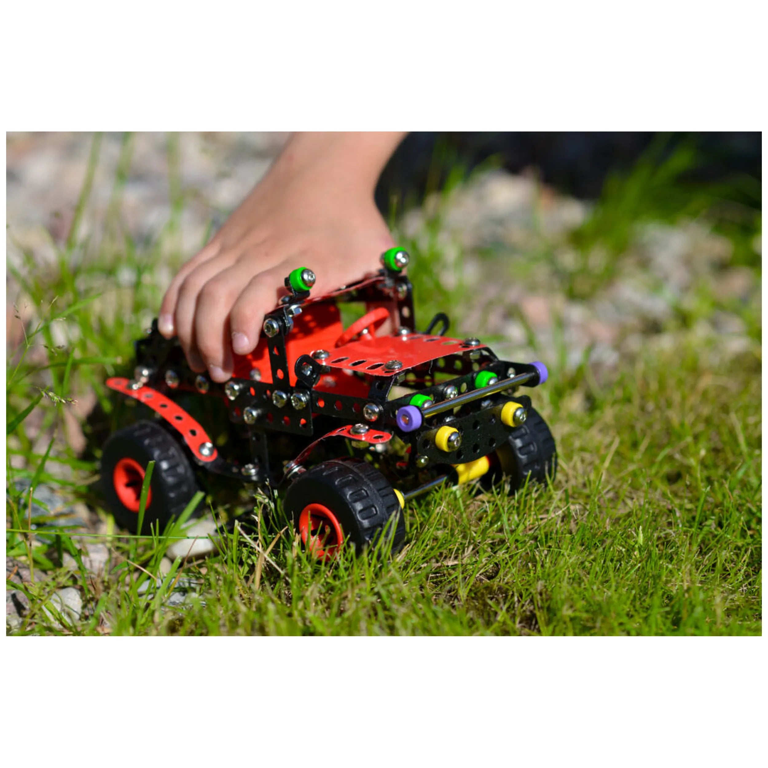 Child's hand building Alexander Black Spider off-road toy on grass, with red and black metal and plastic components.