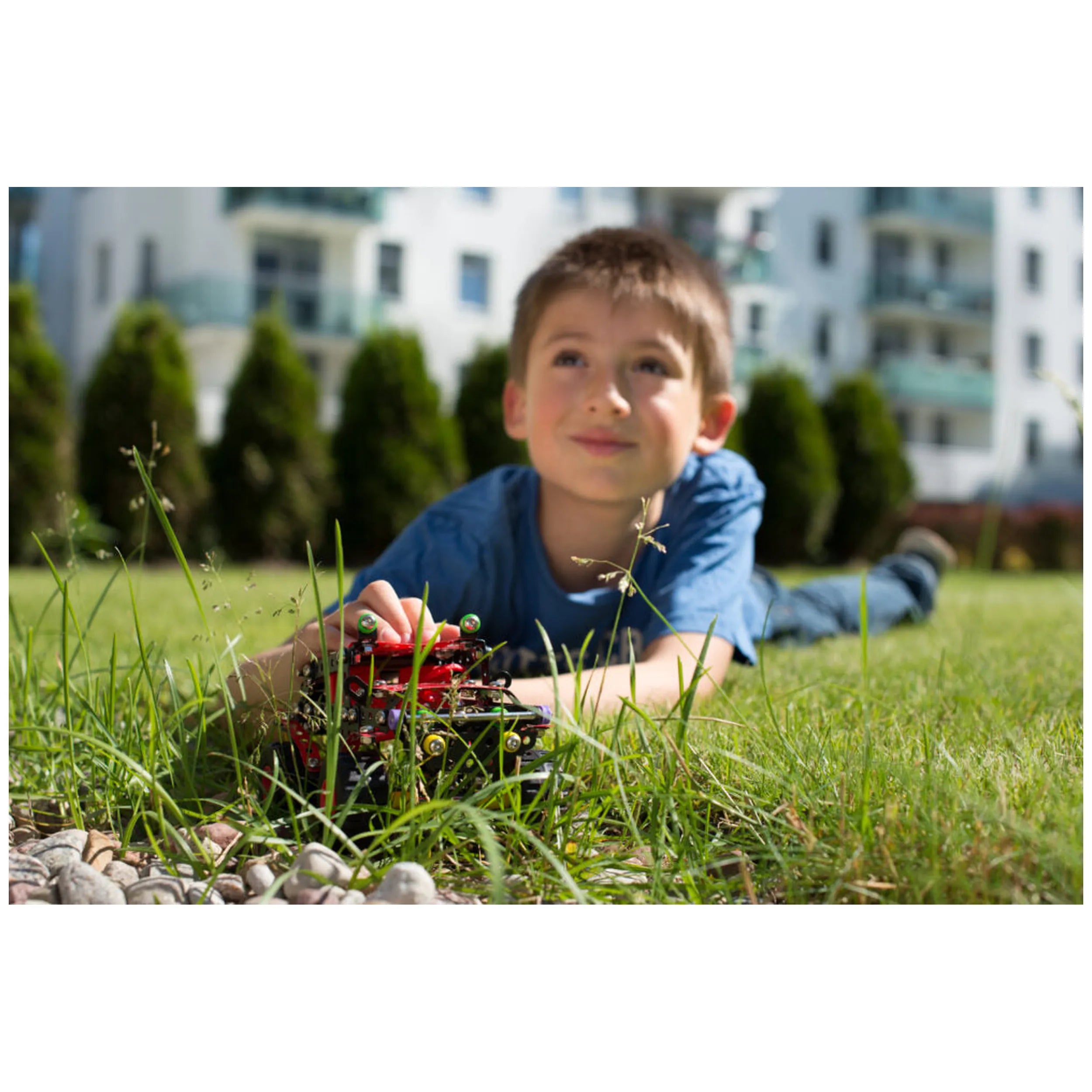 Child playing with Alexander Black Spider construction toy on grass, smiling, with buildings and trees in the background.