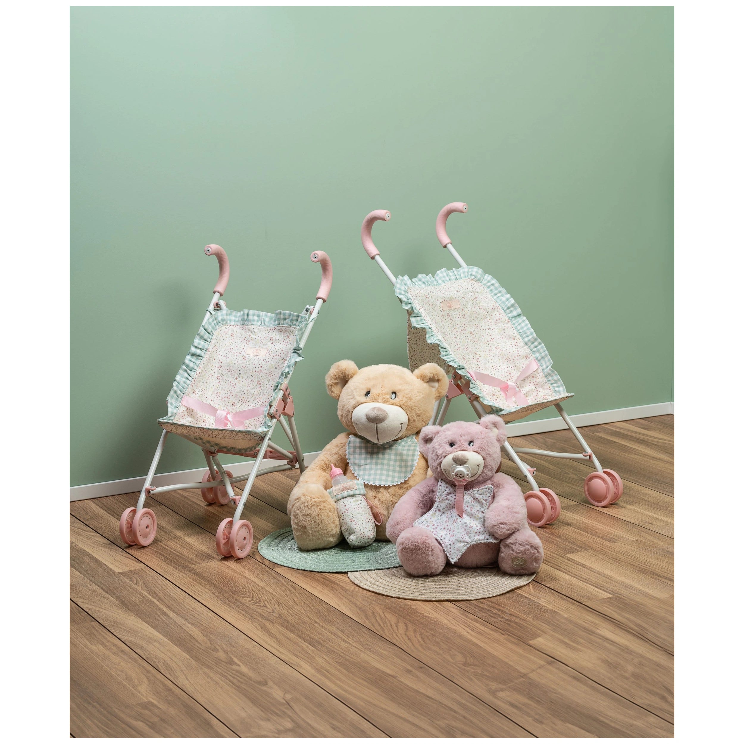 Toy strollers with pink handles beside two plush teddy bears on wooden floor, against a soft green wall.