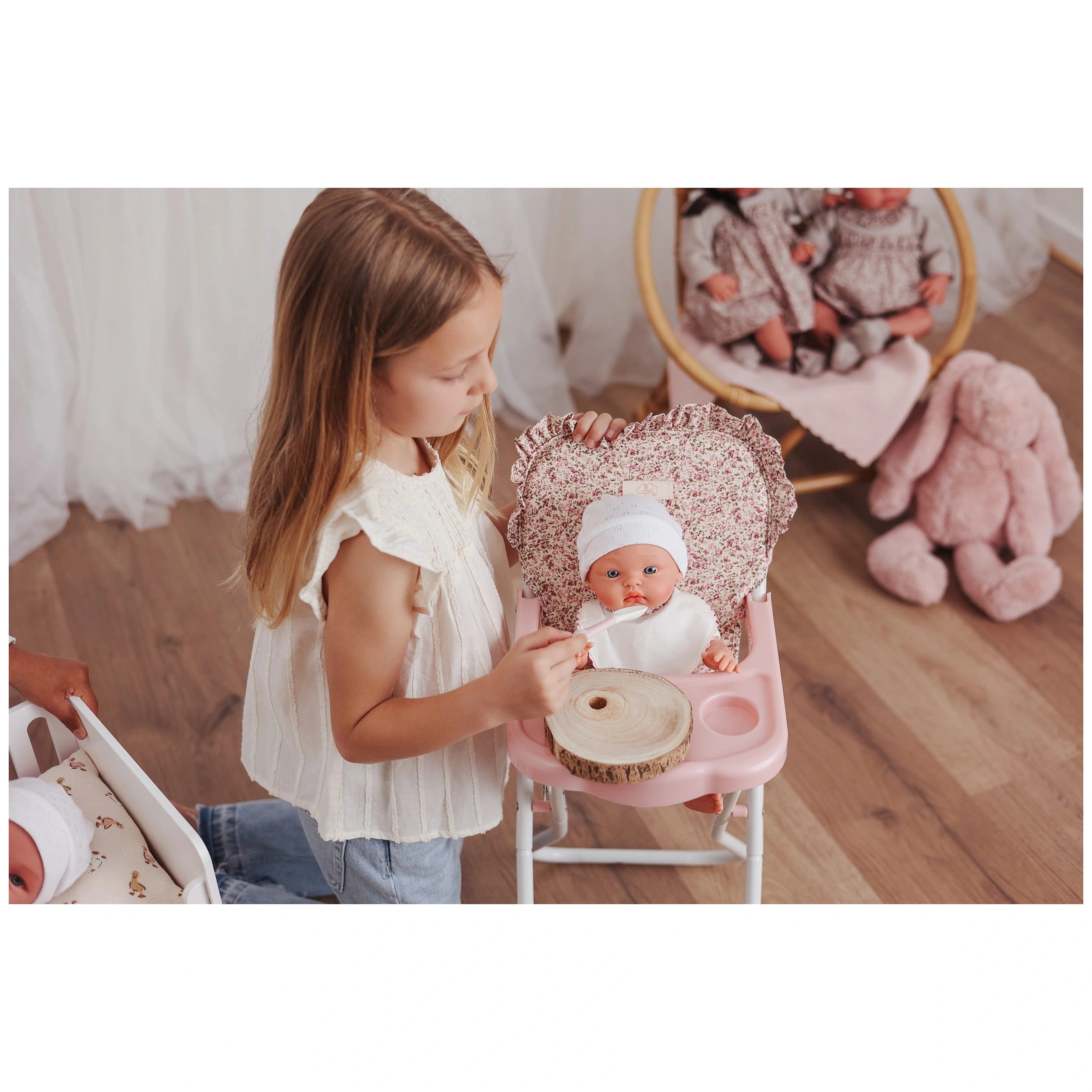 A young girl feeds a doll in a pink high chair with a floral pattern, featuring a sturdy white metal structure and soft ruffled fabric.
