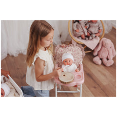 A young girl feeds a doll in a pink high chair with a floral pattern, featuring a sturdy white metal structure and soft ruffled fabric.