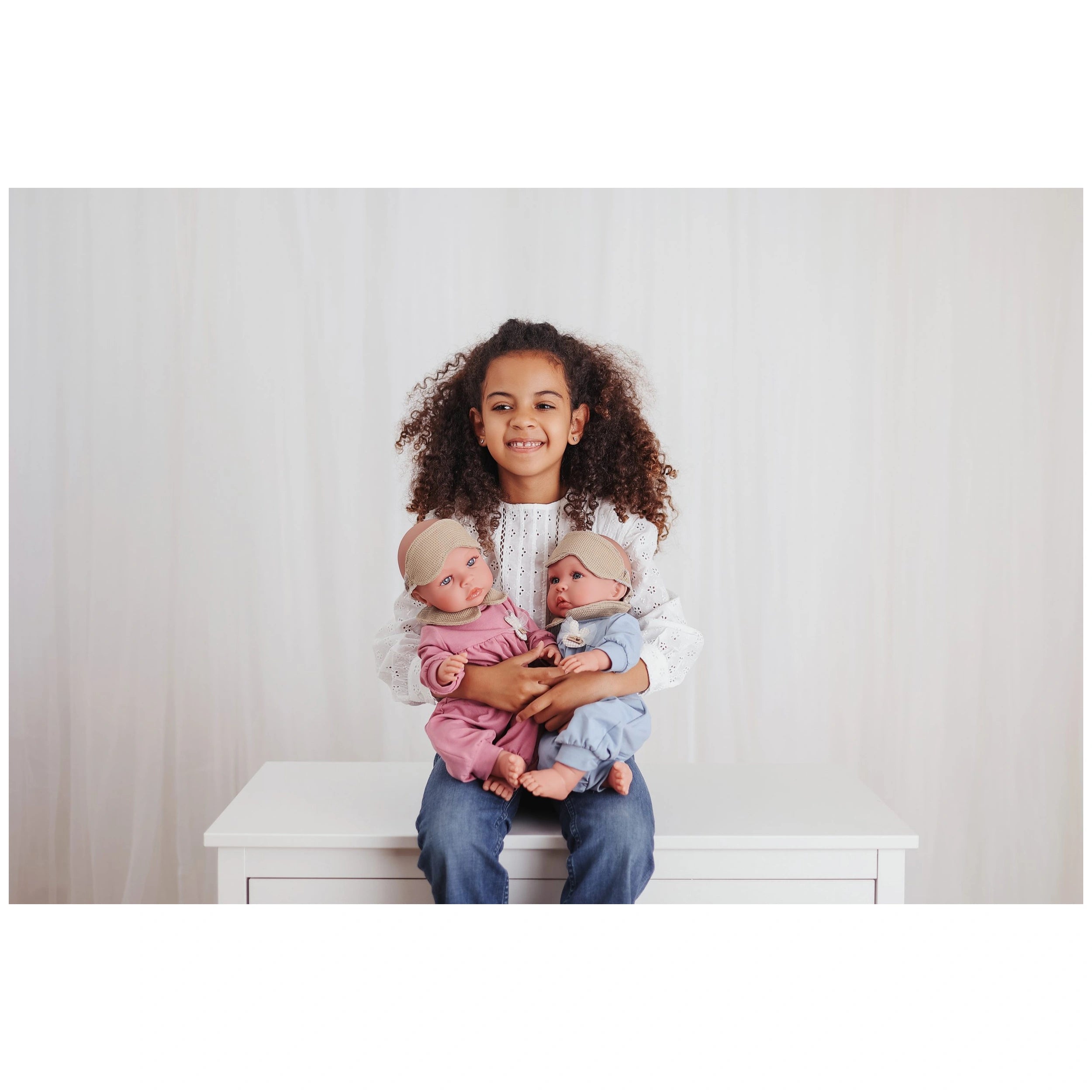 Child holding two realistic dolls, one in pink pajamas, one in blue pajamas, both wearing headbands, seated on a white table.