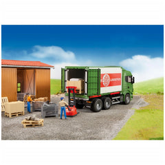 Green toy truck with a cargo container, figures loading pallets and crates, Bruder Logistics set in a rural setting.