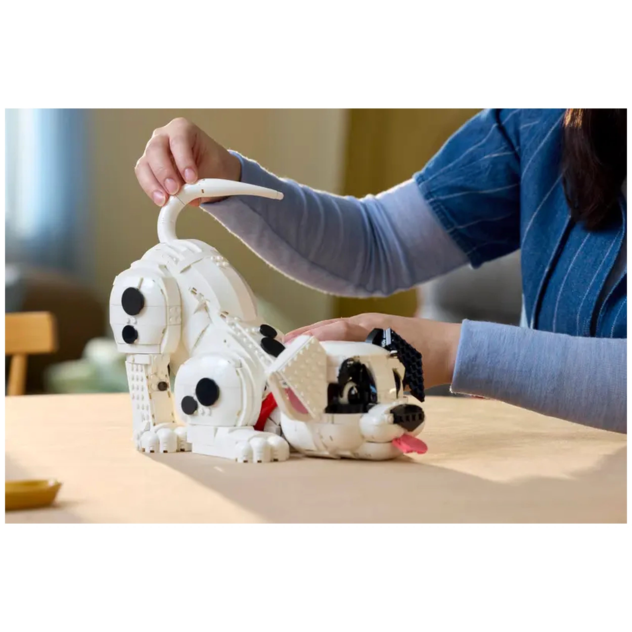 A person is assembling a white Dalmatian puppy build, featuring black spots and a pink tongue, on a wooden table.