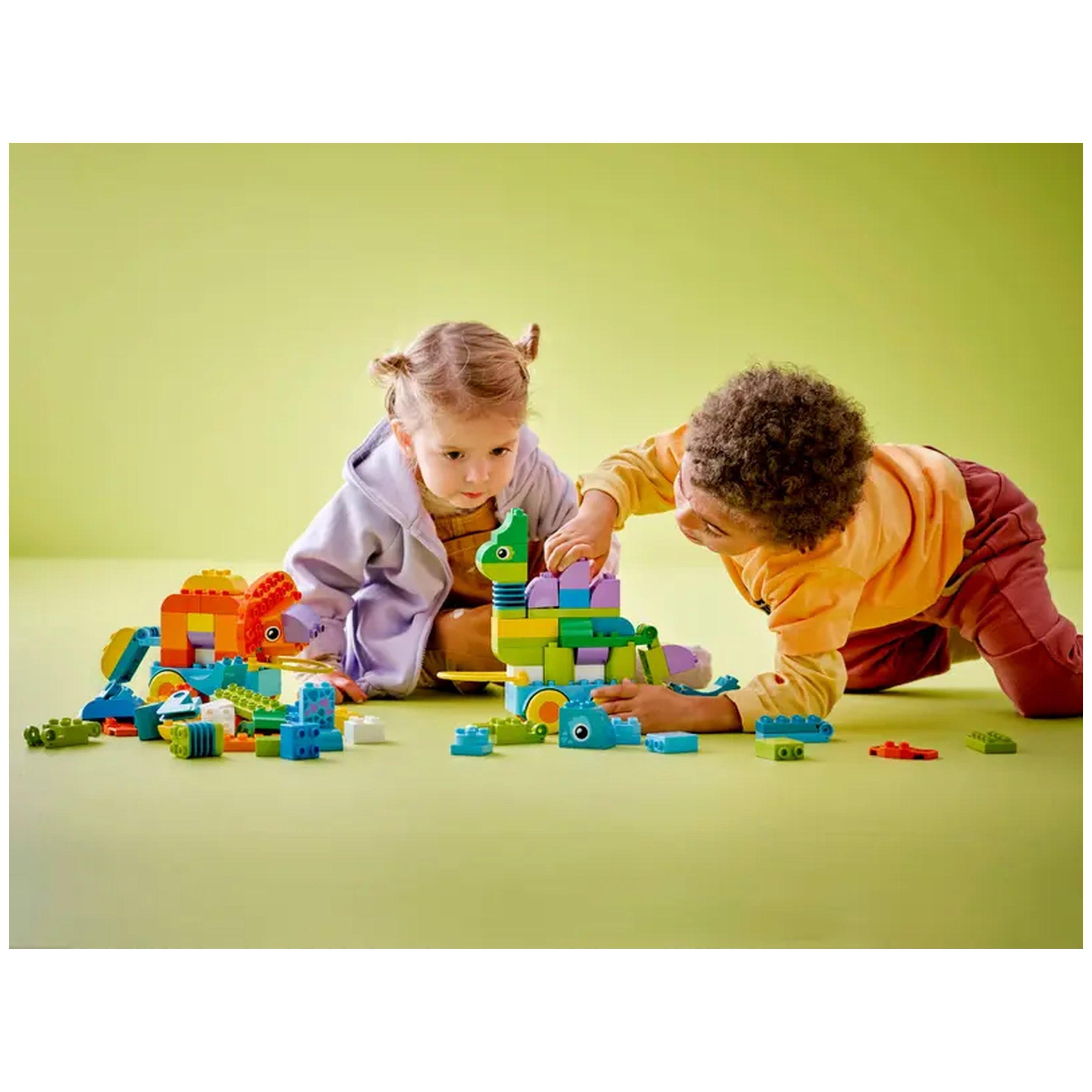 Two children engaged in building with colorful LEGO bricks on a light green floor, surrounded by dinosaur models and various construction pieces.