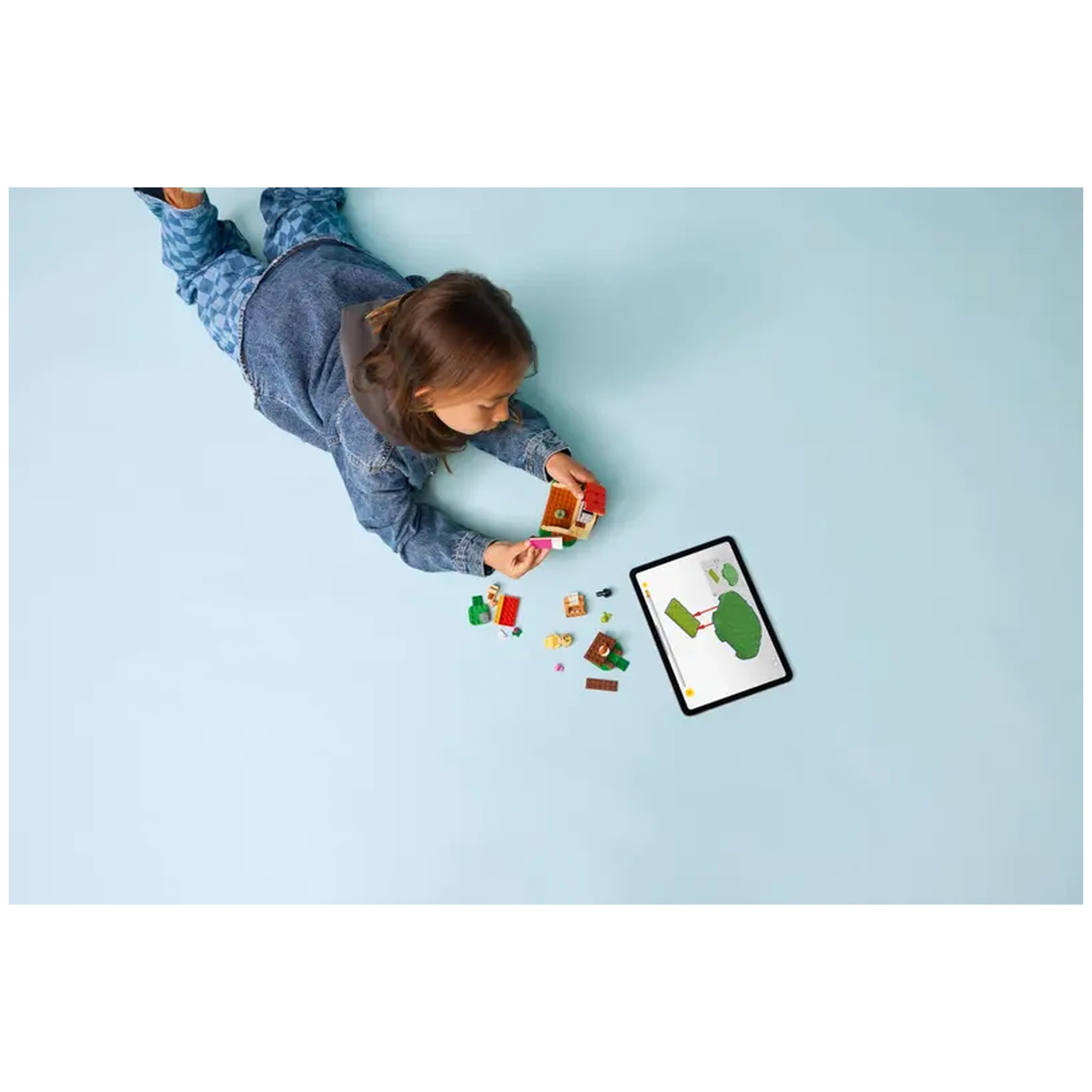 A child lying on a light blue surface assembles LEGO pieces while looking at a tablet showing a construction guide for Goldie's Cozy House.