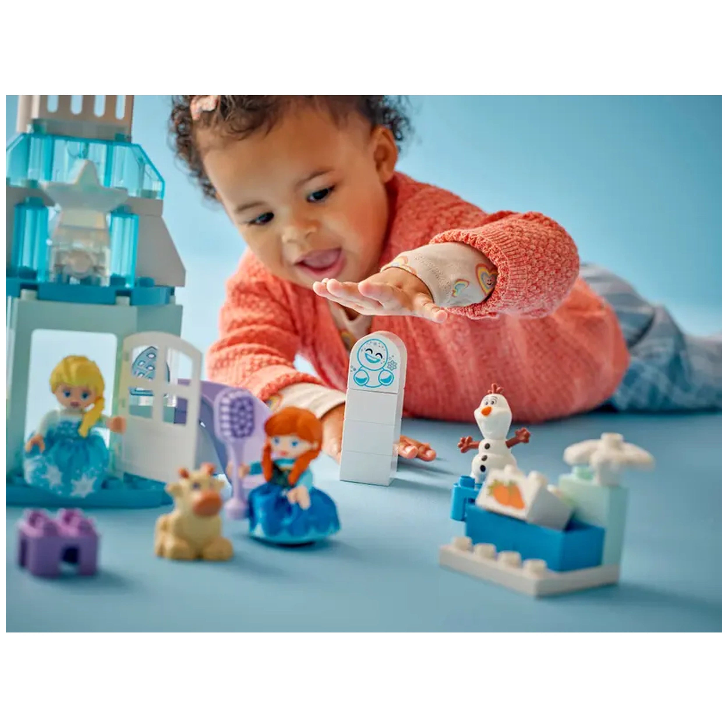A toddler in a coral sweater joyfully reaches for LEGO® DUPLO® characters and a colorful castle set. The scene includes Elsa, Anna, Olaf, and playful accessories on a light blue surface.