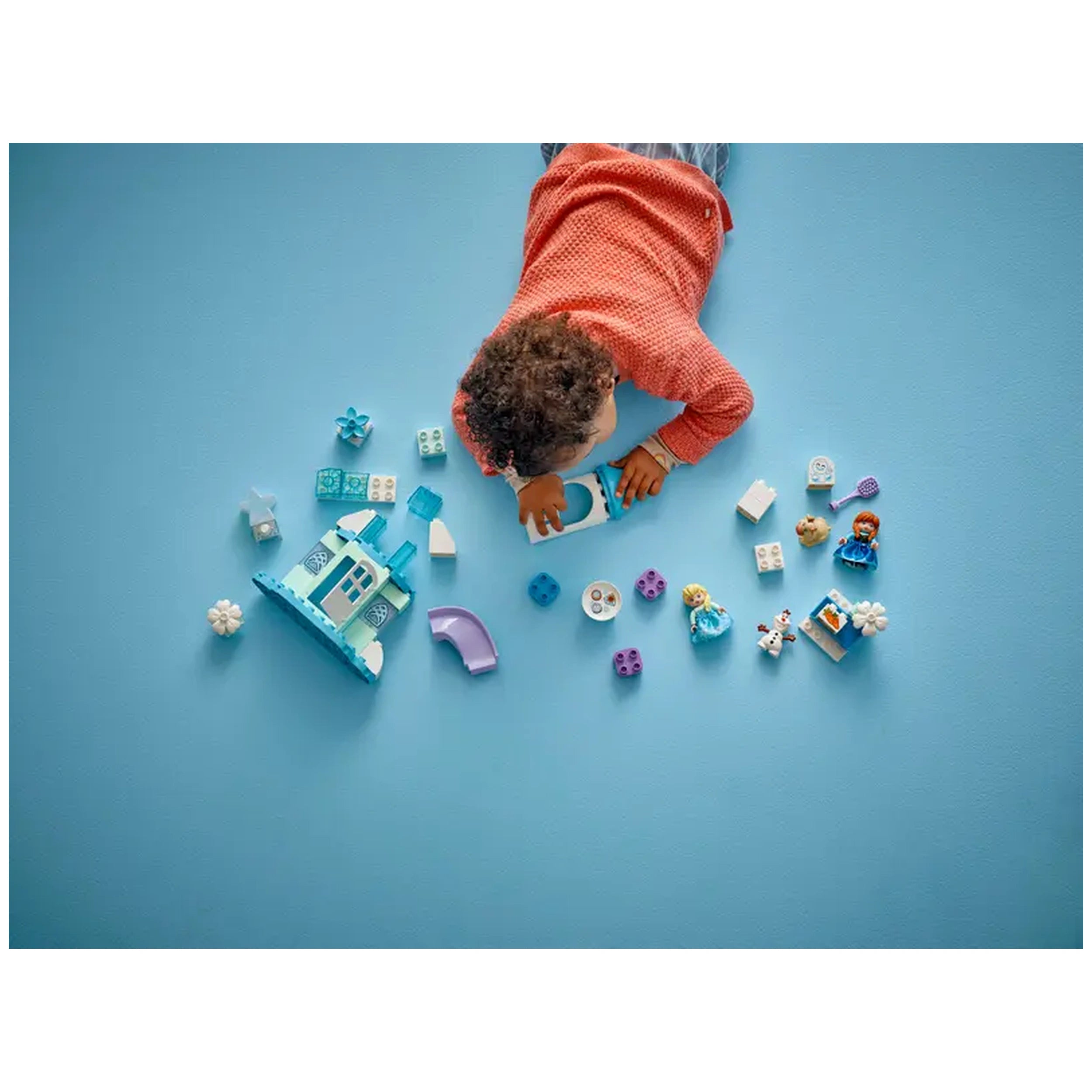 A child in an orange sweater plays on a blue surface, building with LEGO® DUPLO® Frozen characters and accessories, including a castle, Elsa, Anna, and Olaf.