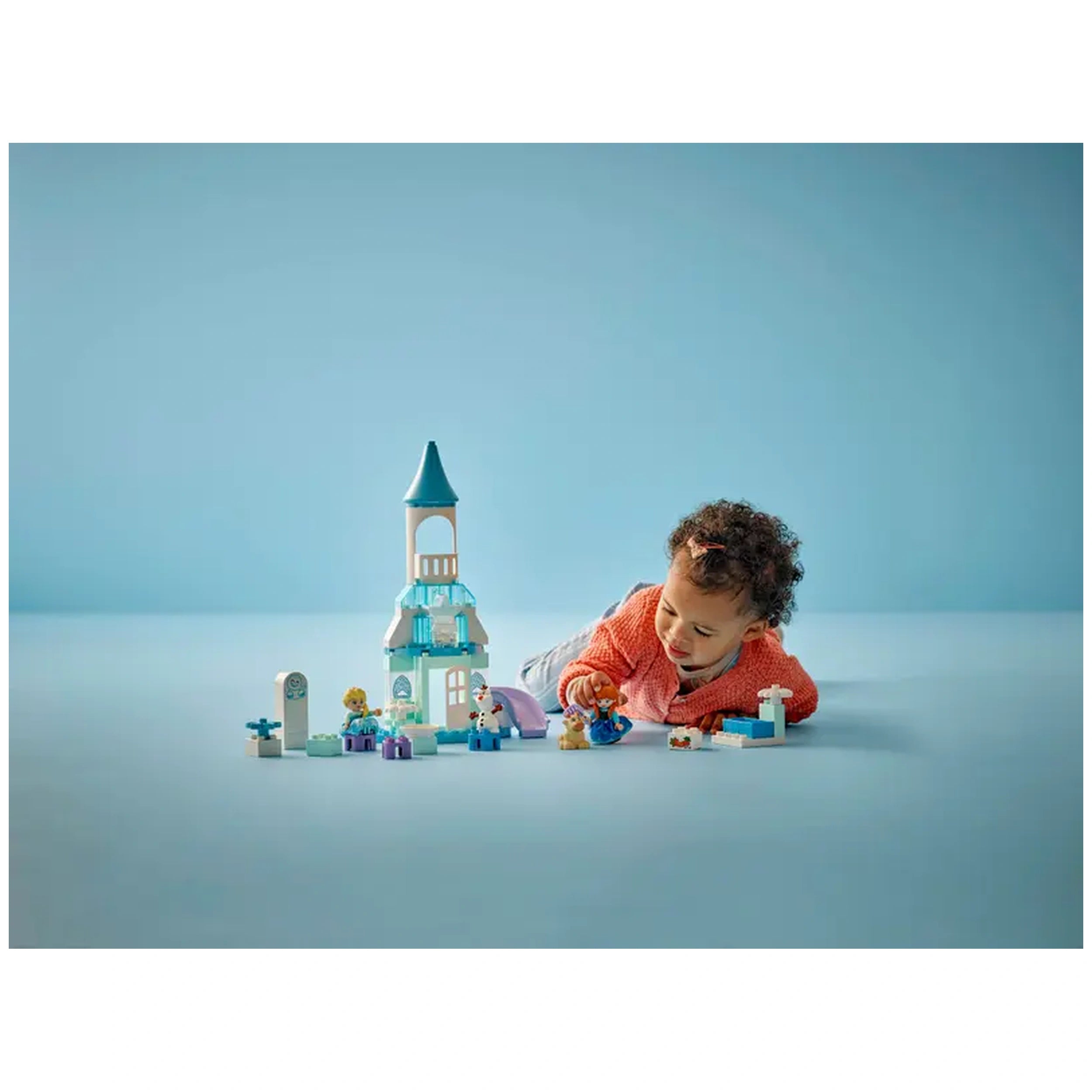 A young child in an orange sweater plays on a light blue surface with a LEGO® DUPLO® Frozen Castle set, featuring colorful blocks and characters.