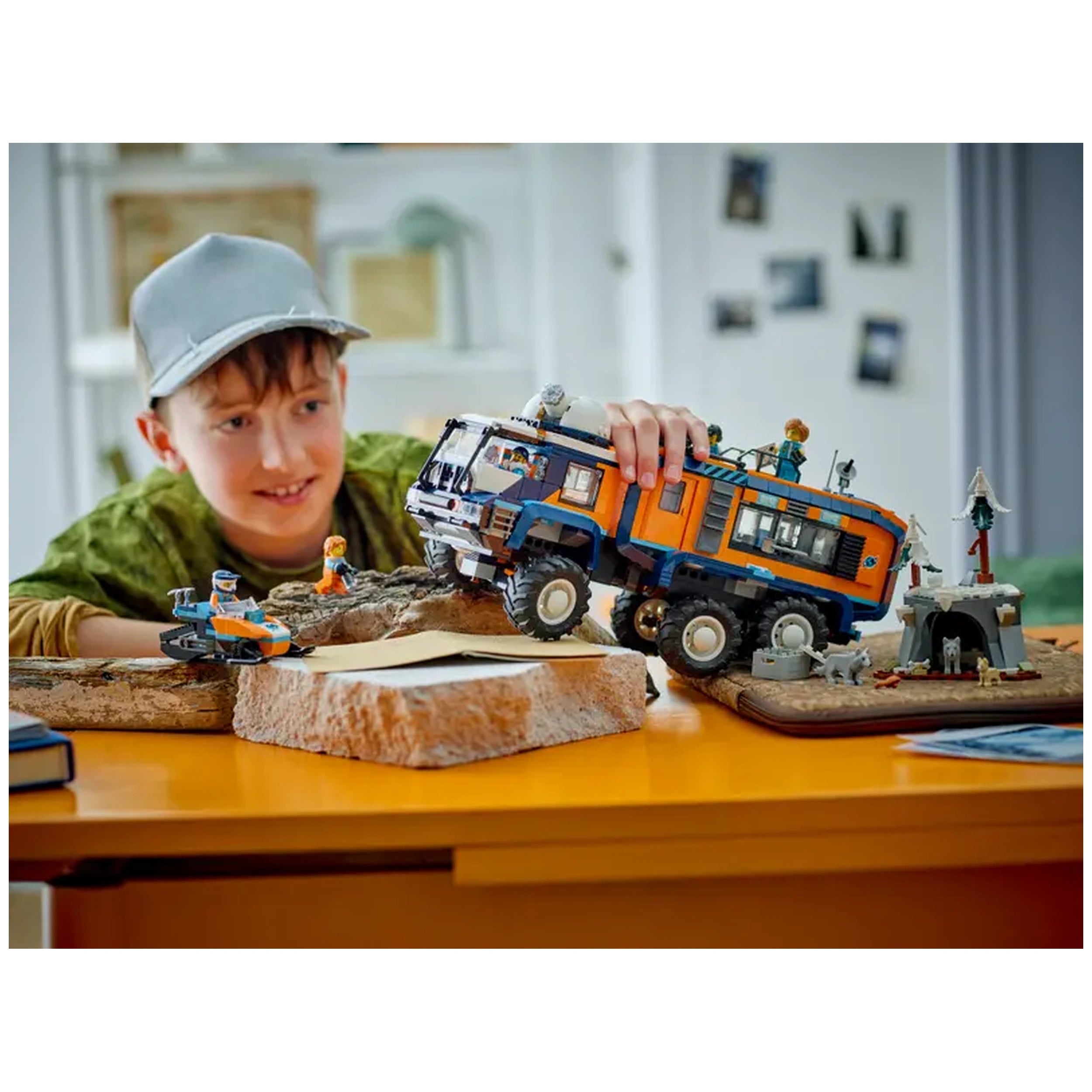 A child wearing a gray cap excitedly maneuvers the colorful LEGO Arctic Explorer Science Lab Truck over rocky terrain on a bright yellow table. Beside the truck, a small snowmobile and an imaginative Arctic scene with mini figures and a makeshift shelter are displayed.