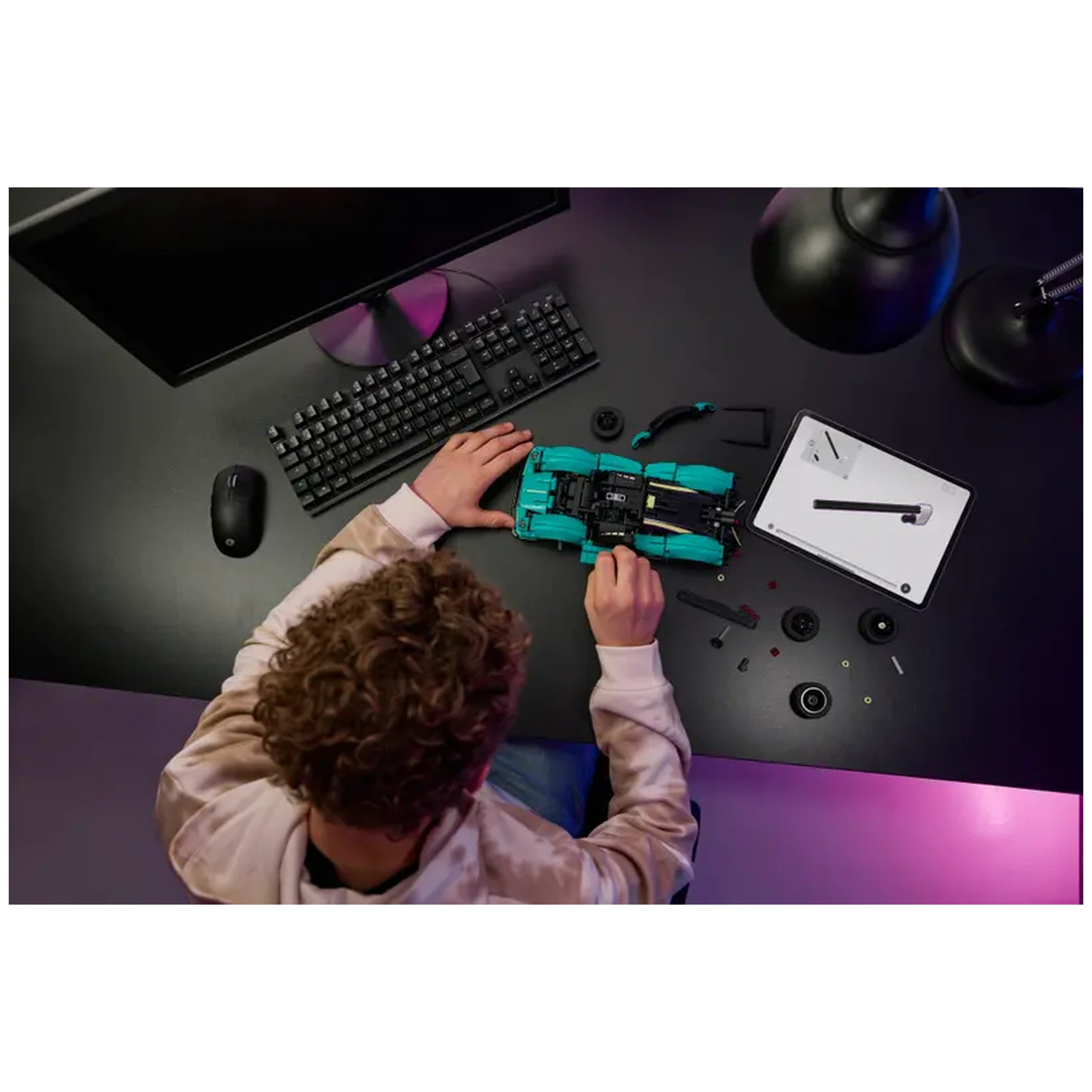 A child assembling the LEGO Aston Martin Valkyrie construction set on a black desk, surrounded by various pieces and a tablet with instructions. A computer and keyboard are visible in the background.