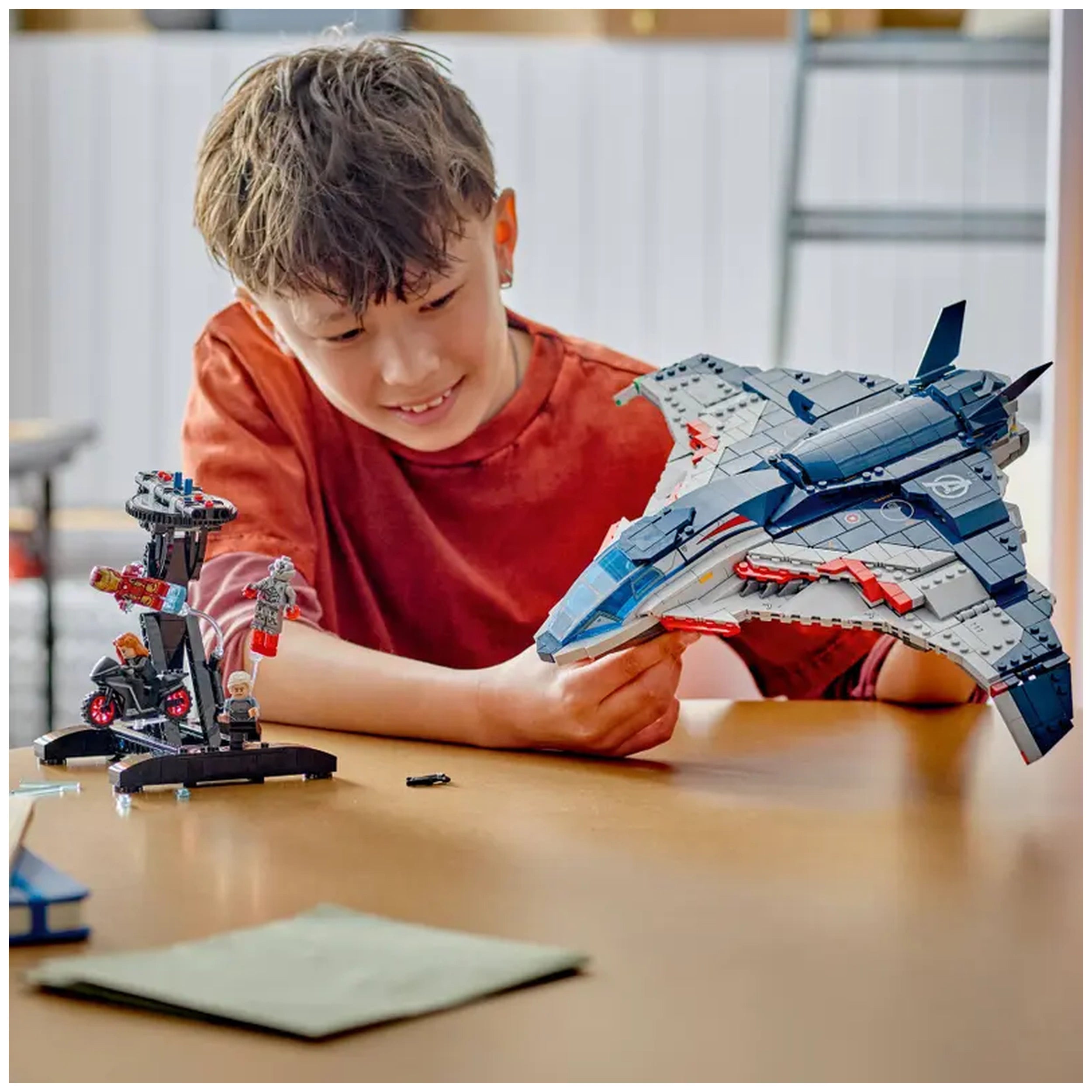 A young boy smiles while holding a LEGO Quinjet model from the Avengers franchise, showcasing its detailed design. In the foreground, a smaller build appears with minifigures and a control tower on a wooden table.