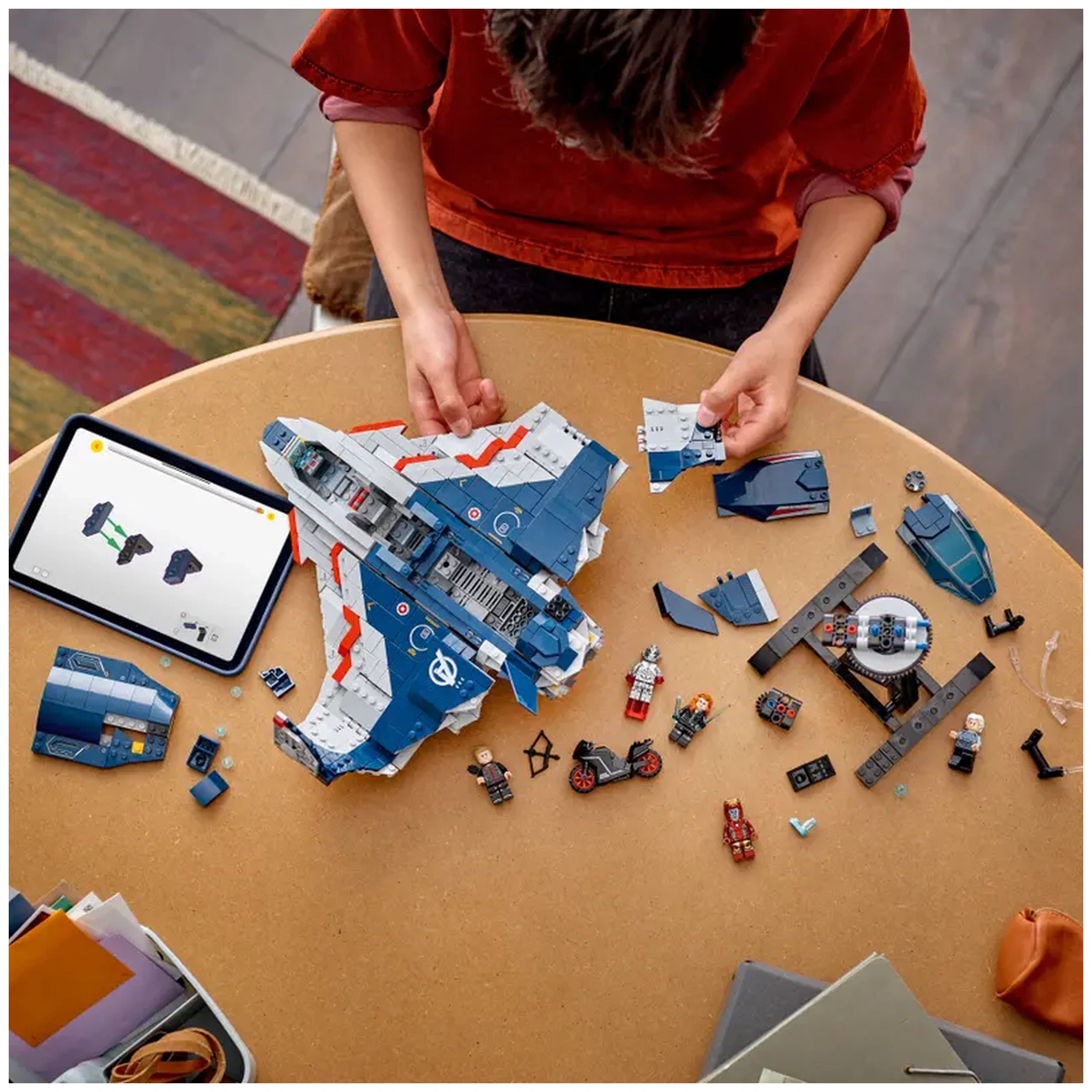 A child in a red shirt builds the LEGO Avengers Quinjet on a round table, surrounded by various pieces, including minifigures and parts. An instruction tablet displays the assembly steps beside them.