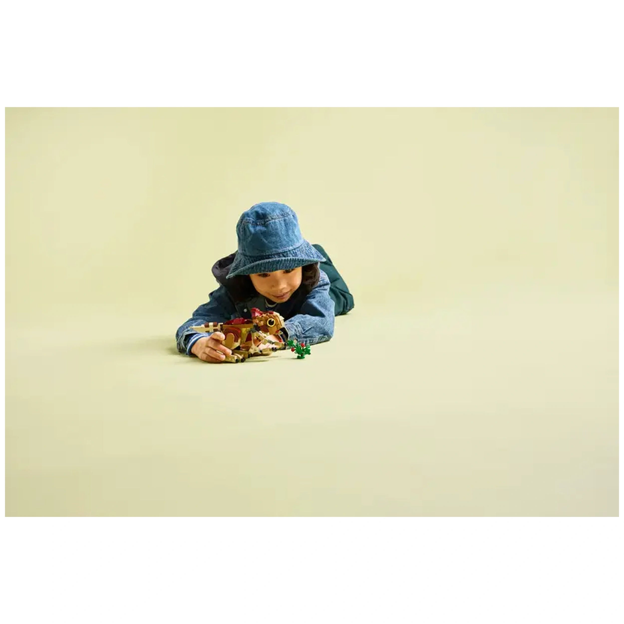 A child wearing a denim jacket and blue bucket hat lies on a light yellow background, playing with the LEGO Baby Dinosaur Dolores set and colorful plant pieces.