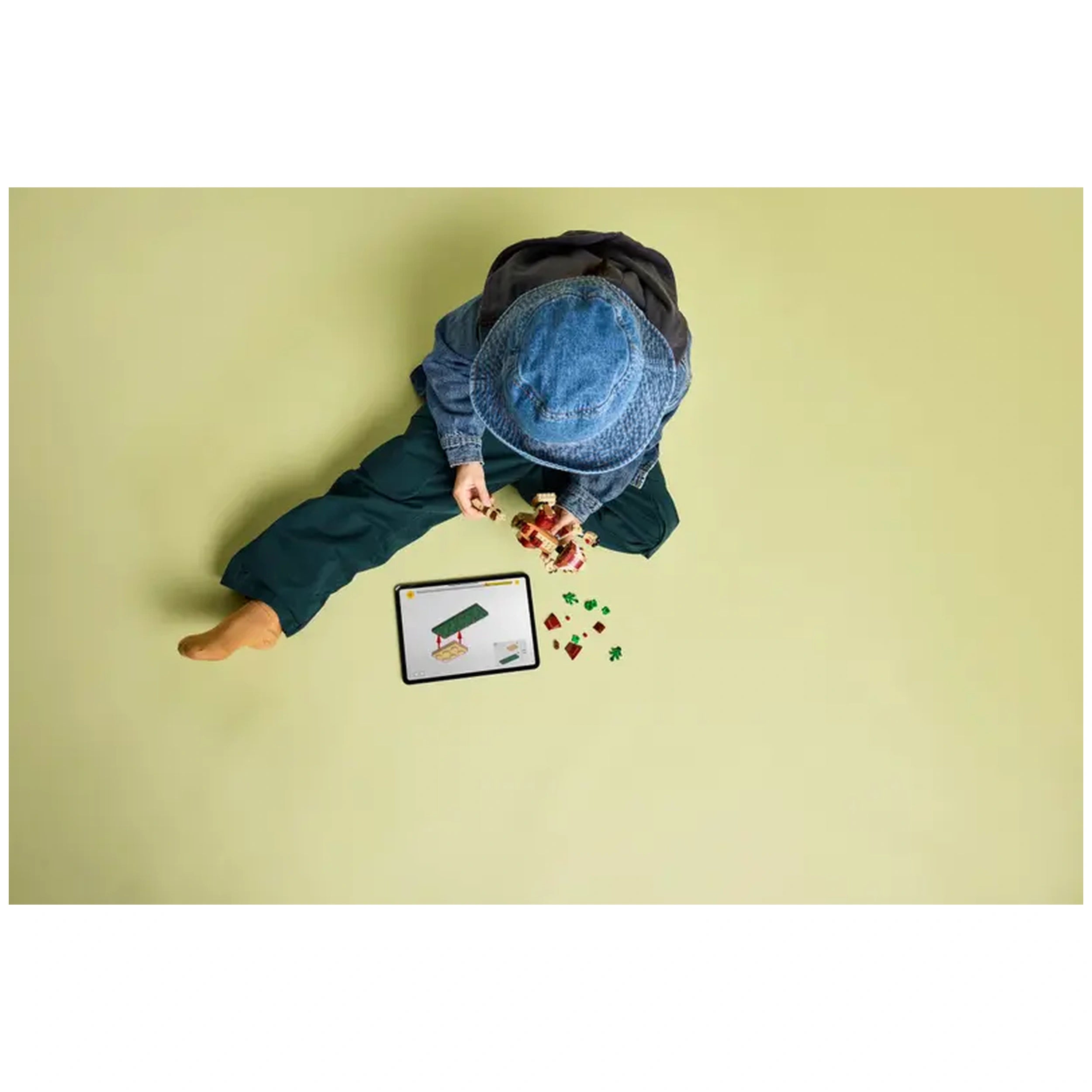 A young child sits on a light green floor, wearing a denim hat and jacket, assembling LEGO pieces. An interactive tablet displays building instructions, surrounded by colorful bricks.