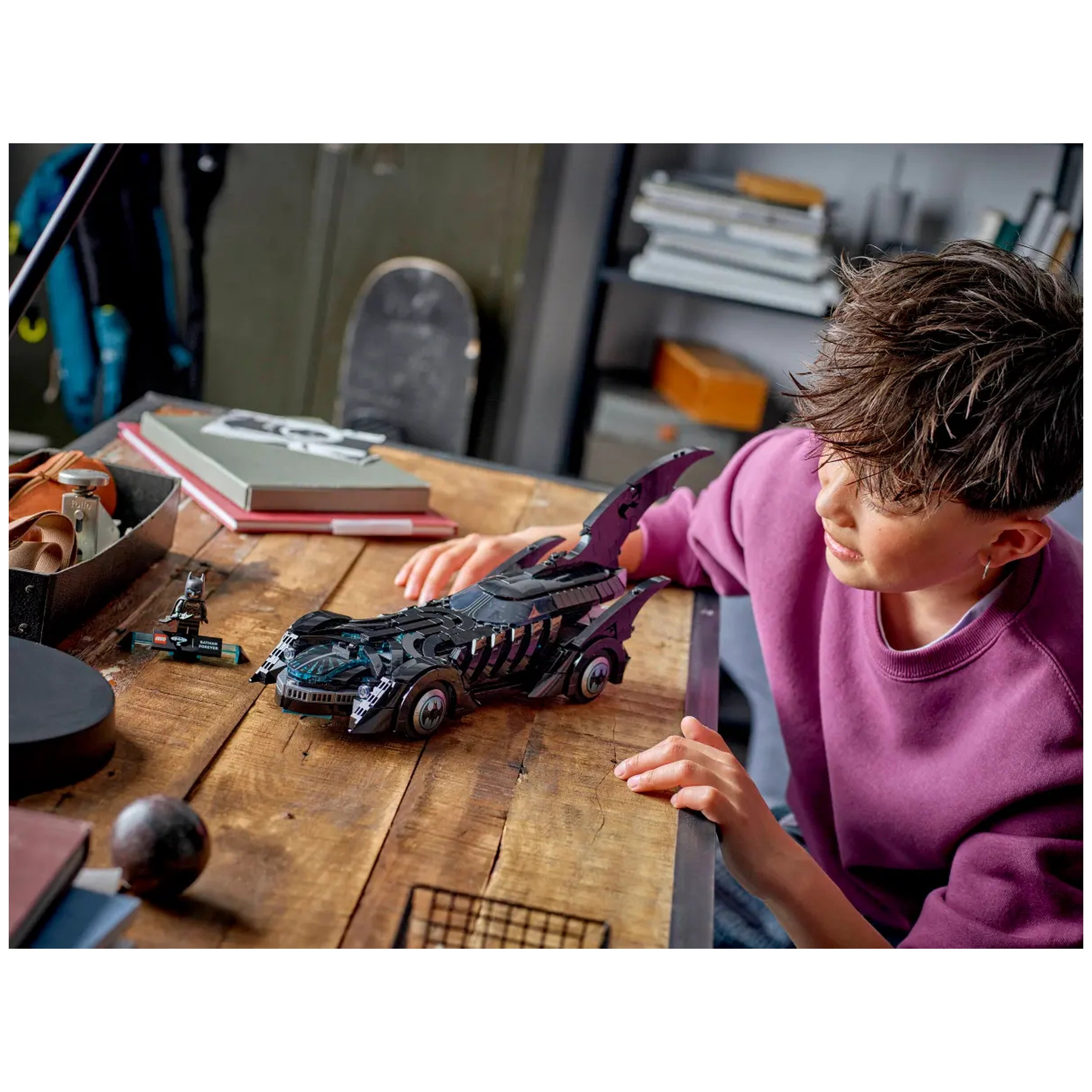 A child in a purple sweater examines the LEGO Batman Forever Batmobile on a wooden table, surrounded by books and a small Batman figure.