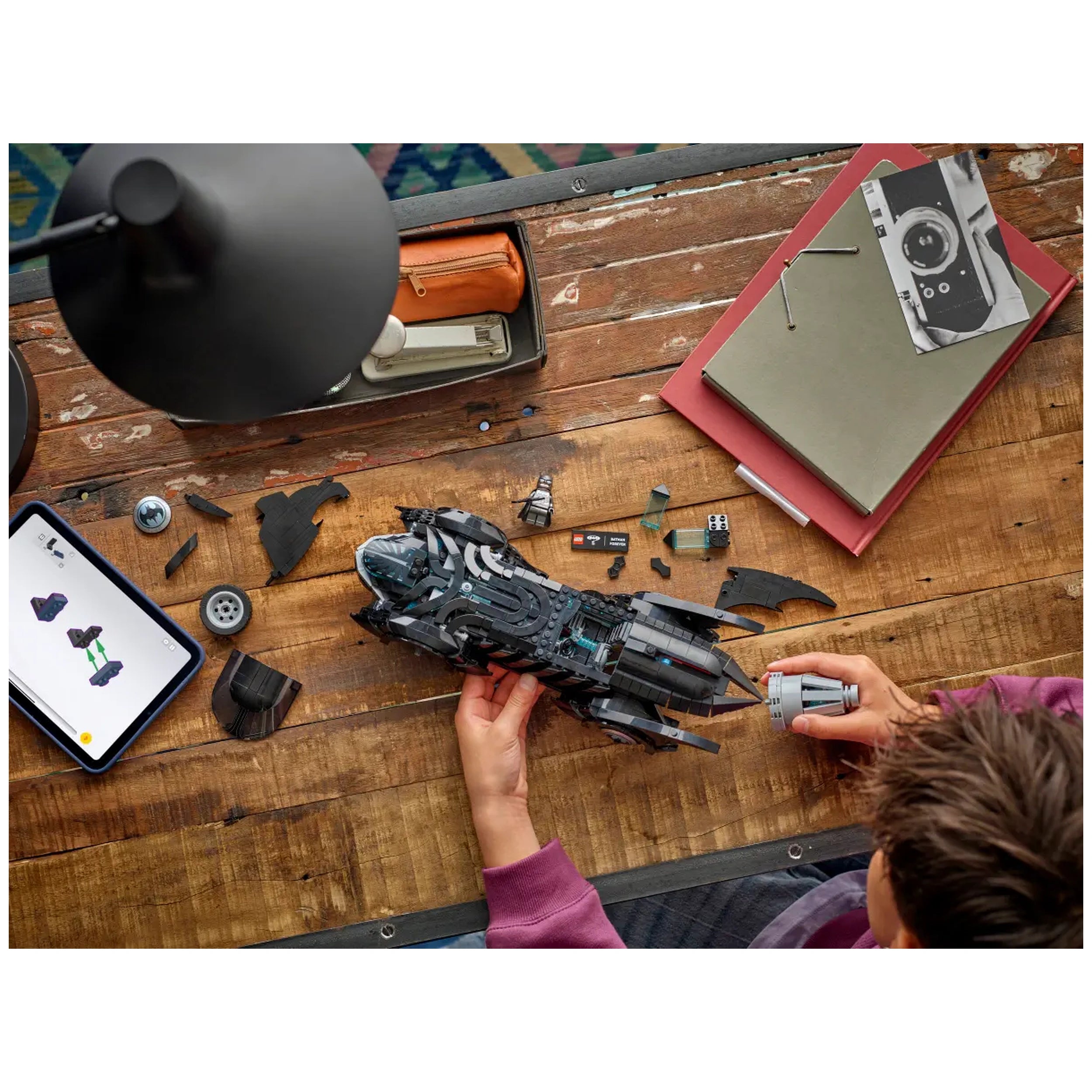 A child assembles the intricately designed LEGO Batman Forever Batmobile on a wooden table, surrounded by building pieces and an instruction tablet.