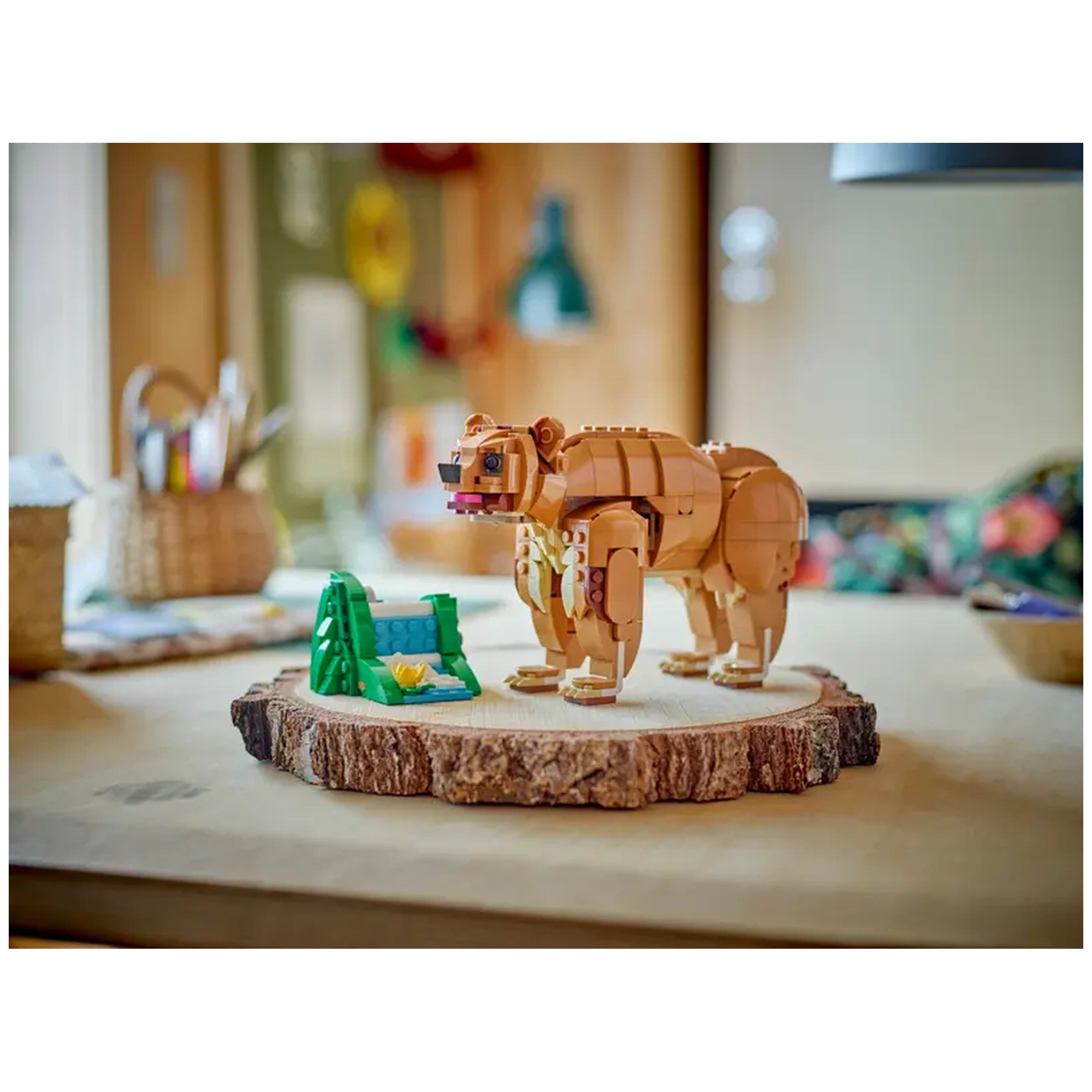 Posable LEGO bear figure in warm brown color, standing on a wooden slice base next to a green tent and yellow flower, with a cozy indoor background.