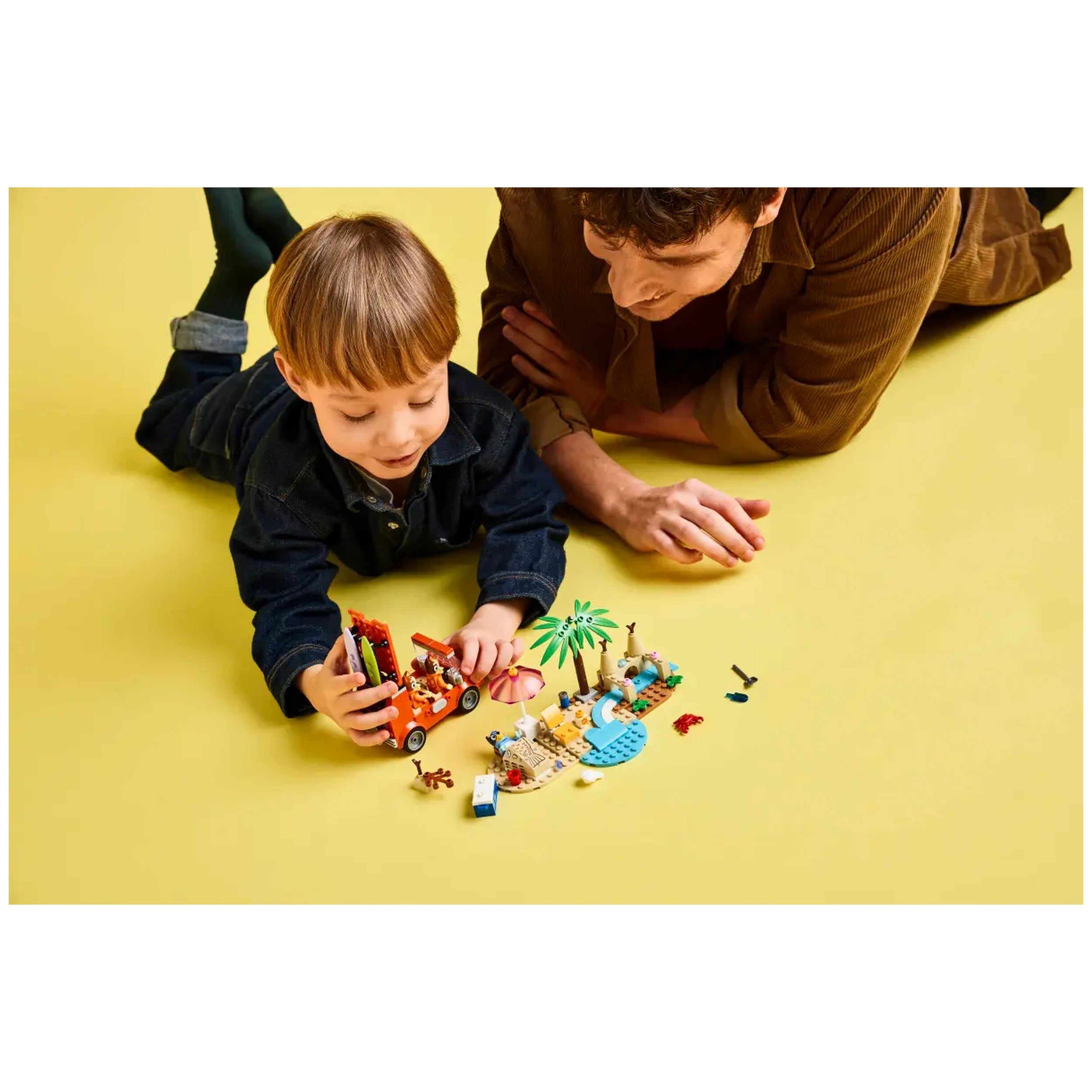A child plays with LEGO's Bluey's Beach & Family Car Trip set on a yellow background, surrounded by colorful toy figures and accessories. A parent watches closely, engaging in imaginative play.