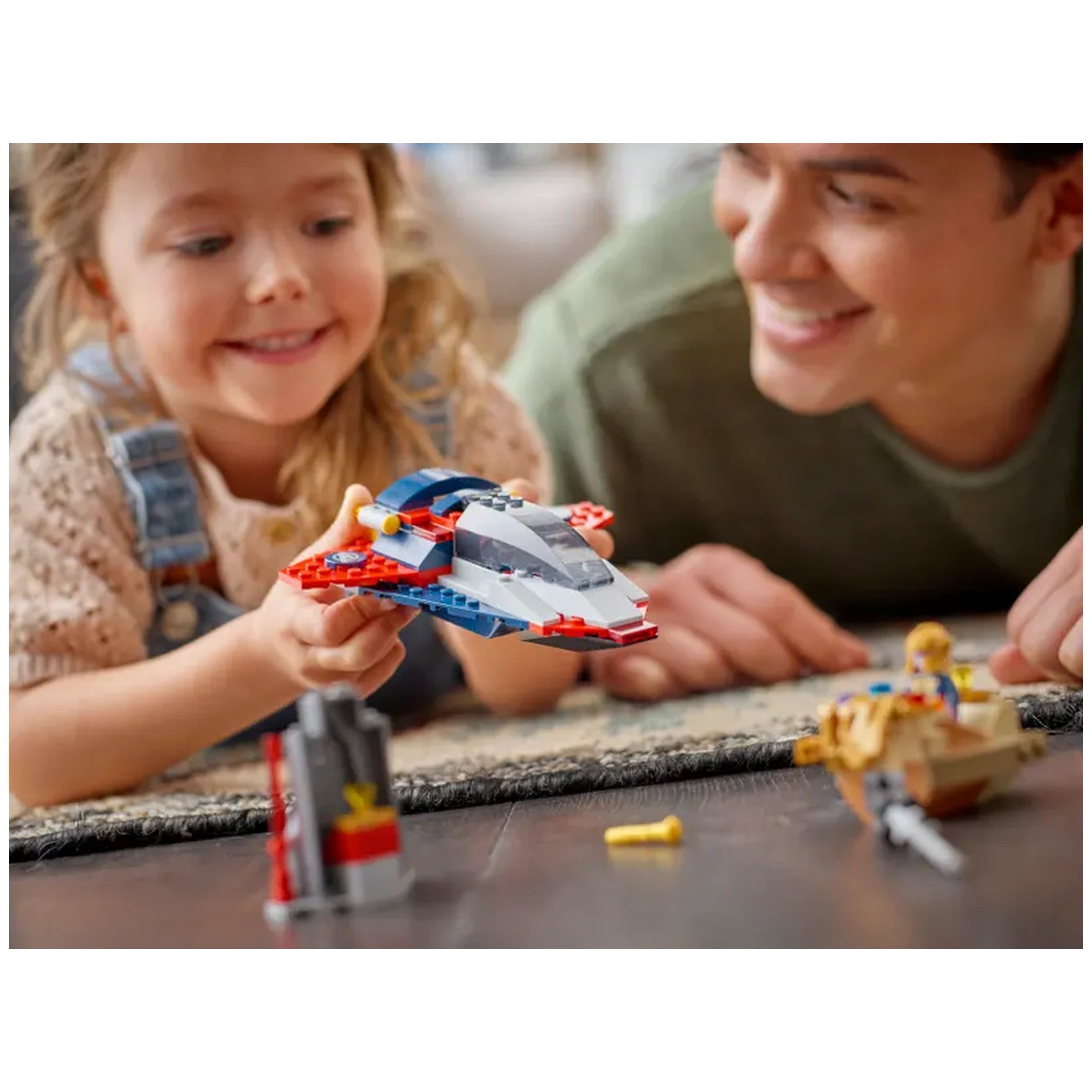 A young girl smiles while holding a colorful LEGO spaceship, with an adult nearby, both engaged in playful creativity on a soft rug.