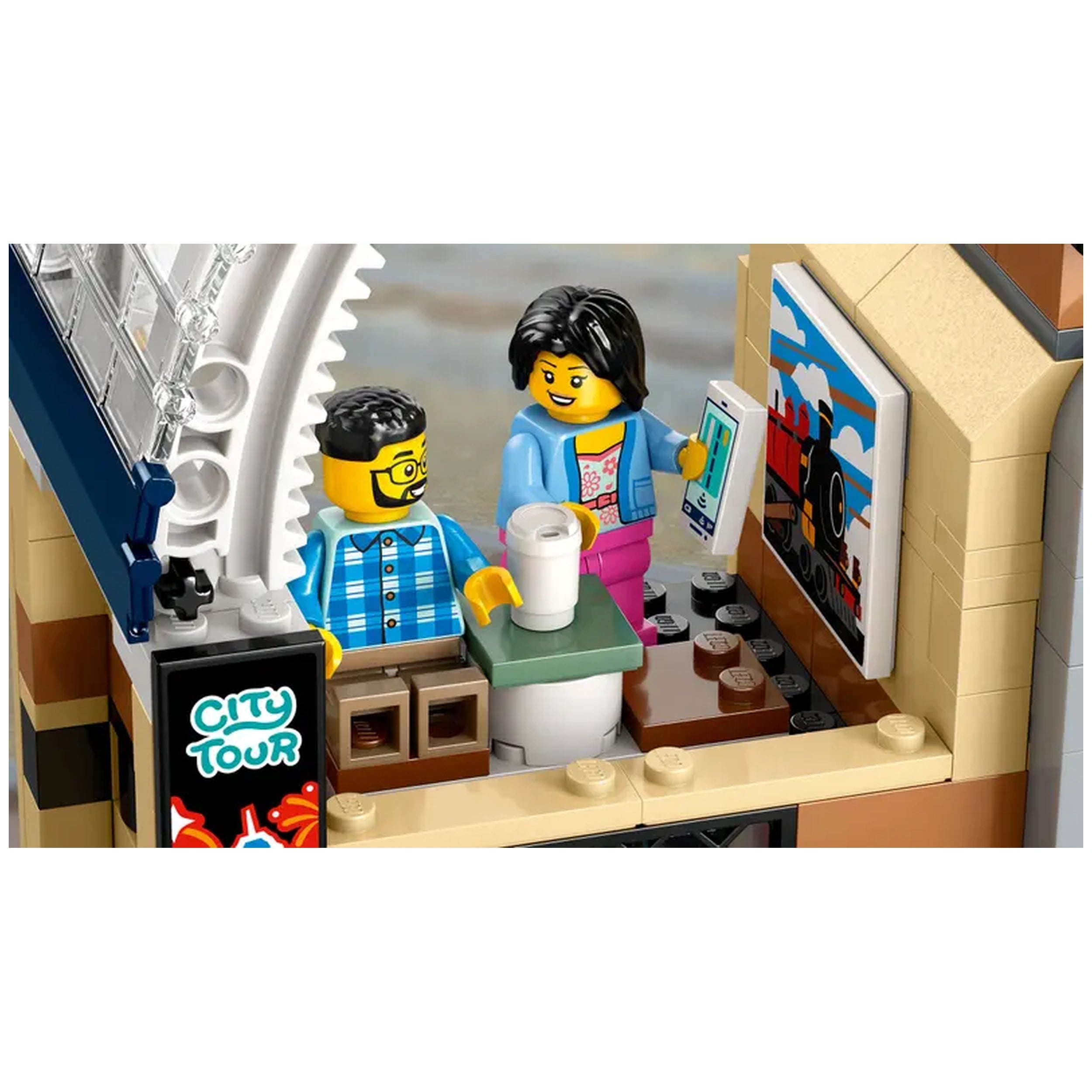 Two LEGO figures interact inside a colorful train station café, one holding a coffee cup and the other a tablet, with a train-themed poster on the wall.