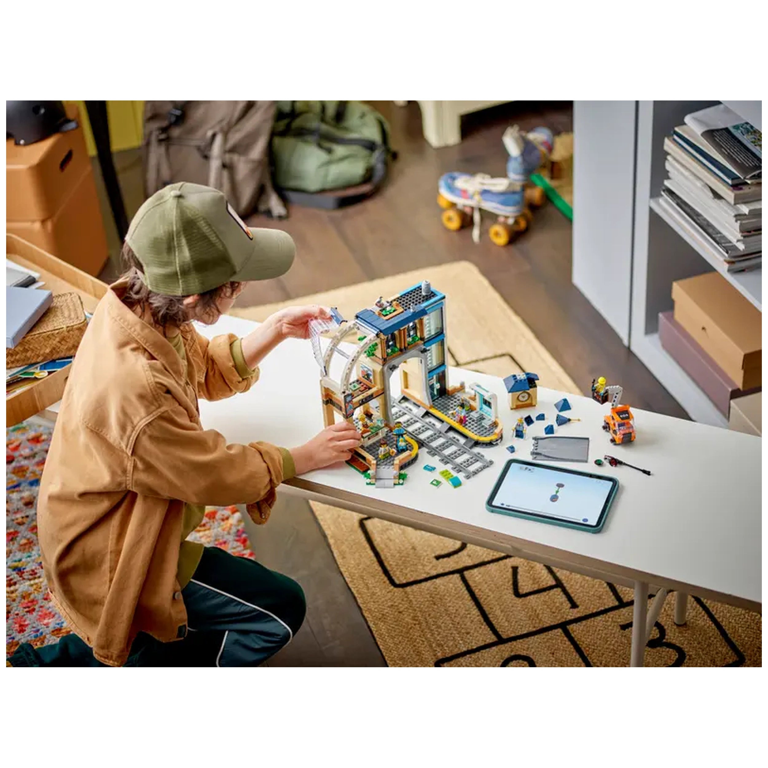 A child builds the LEGO City Central Train Station on a white table, surrounded by colorful blocks and a tablet, in a cozy room.