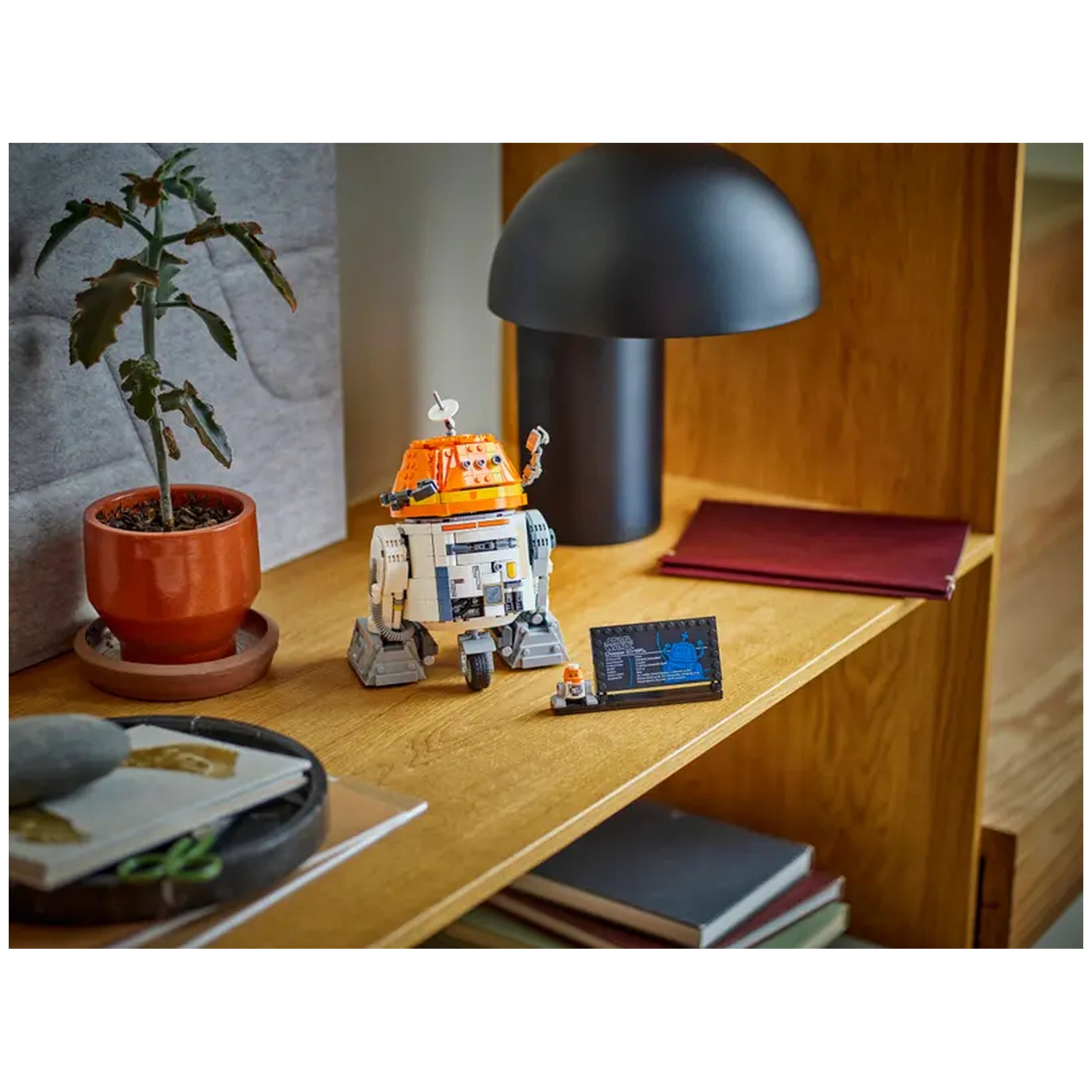 LEGO Chopper construction set displayed on a wooden shelf alongside a potted plant, a lamp, and books, showcasing its vibrant orange details.