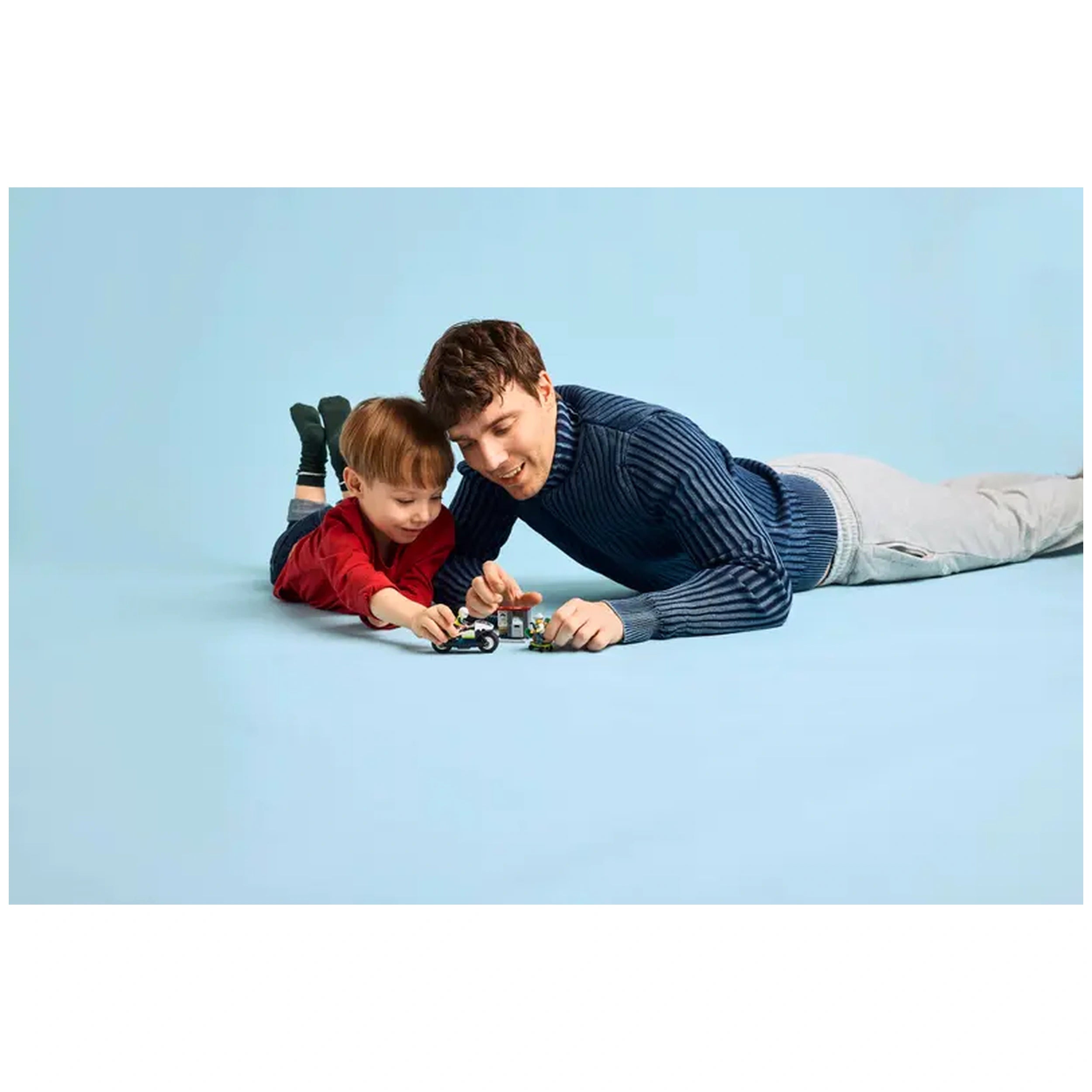 A young boy in a red sweater and a man in a blue sweater lie on a light blue background, focusing intently on a LEGO police motorcycle and crook figure as they play together.