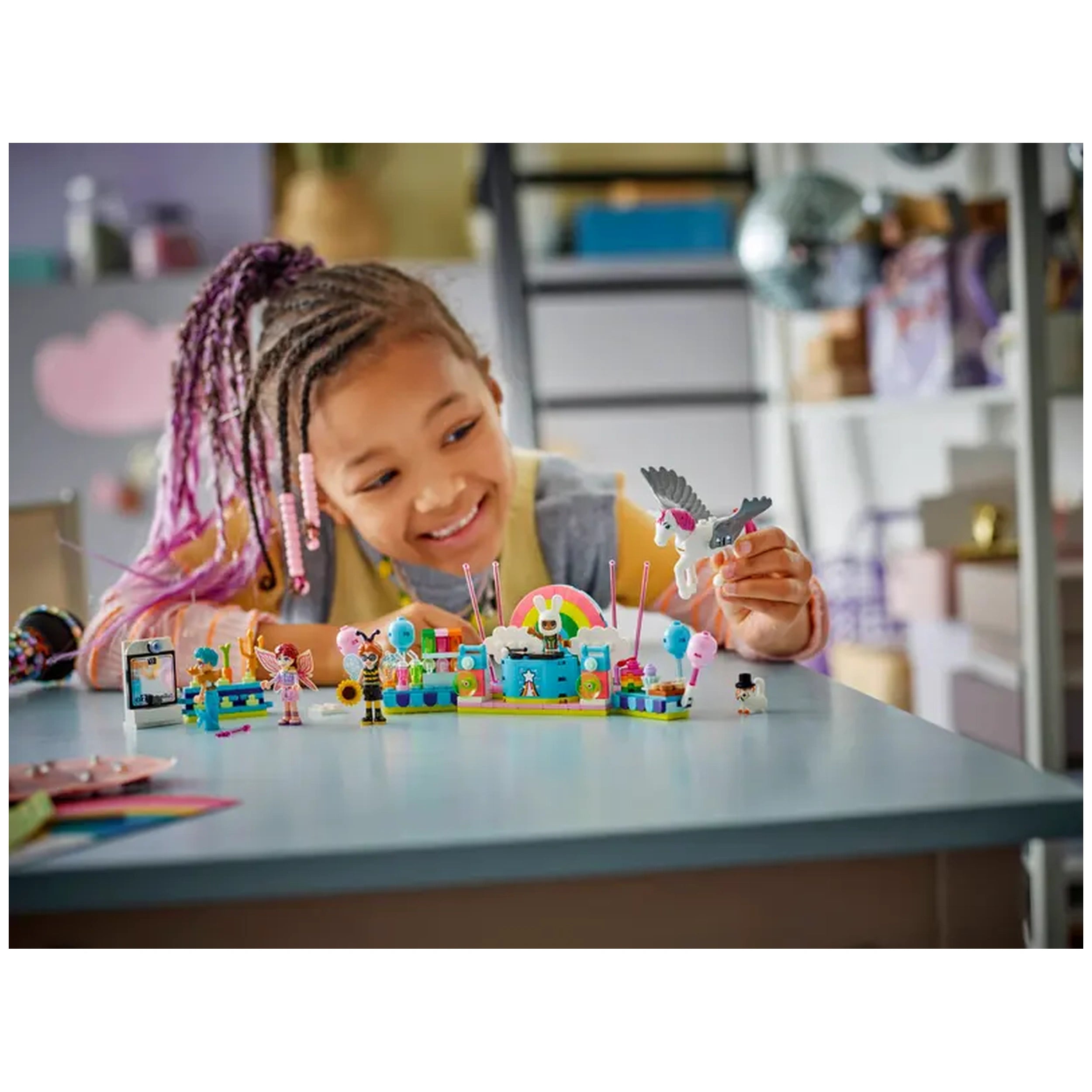 Young child with braided hair and colorful beads smiles while playing with the LEGO® Friends Costume Party set. The vibrant scene includes minidolls, a unicorn, and decorative party elements on a blue table.