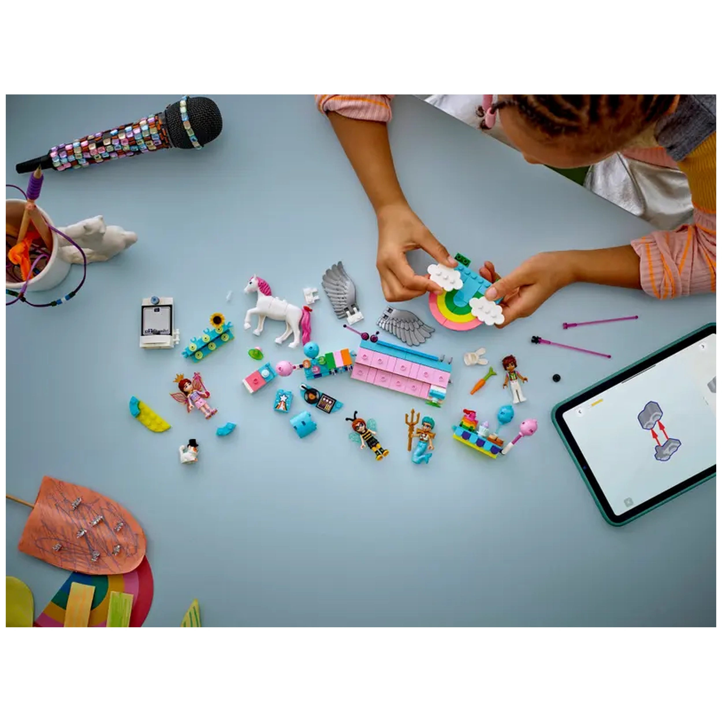 A child's hand holds a colorful LEGO structure featuring a rainbow and clouds, surrounded by vibrant LEGO pieces, minidolls, a unicorn, and a tablet displaying building instructions. A black microphone and various crafting materials are in the background.