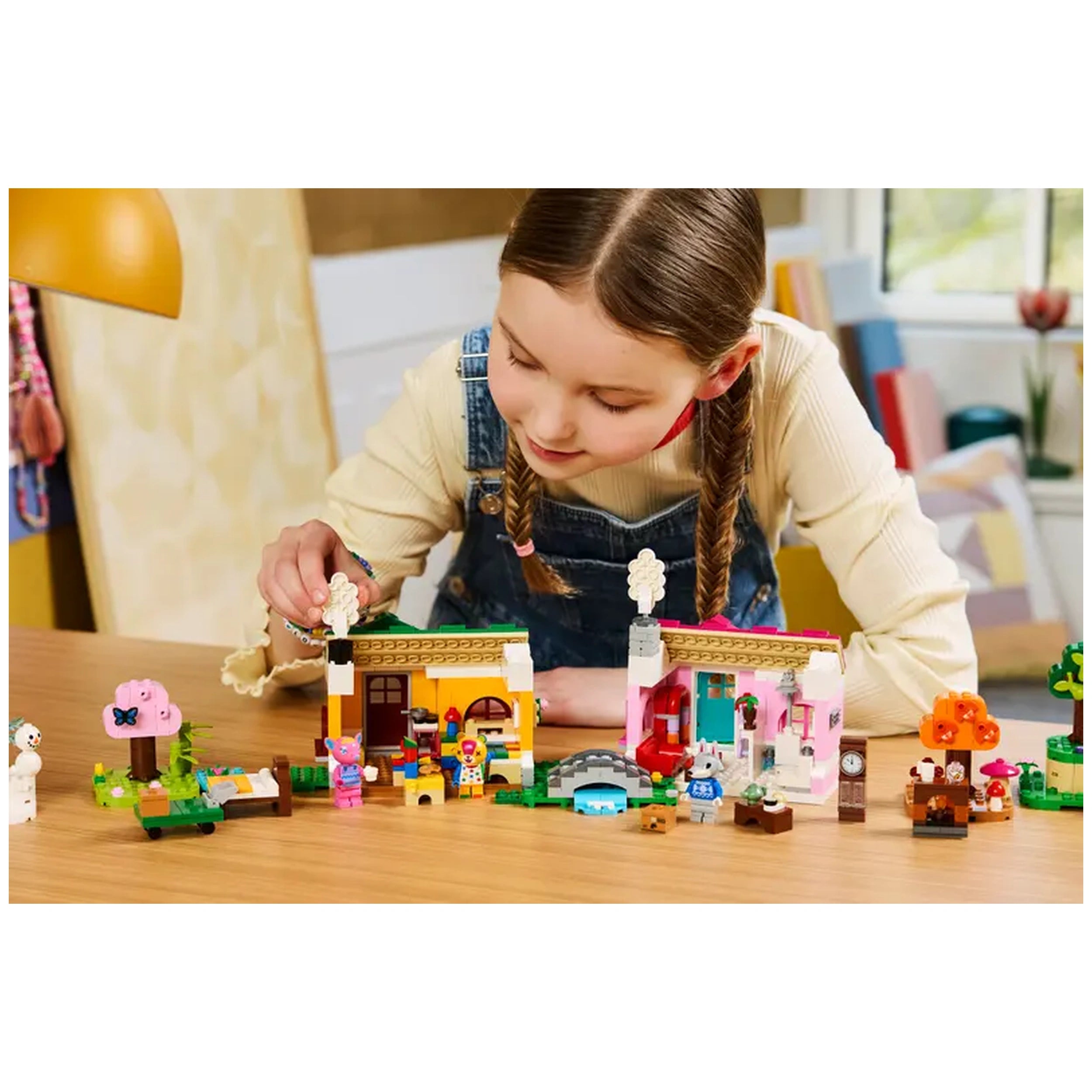 A girl with braided hair carefully arranges a colorful LEGO set depicting two charming houses, surrounded by playful accessories like trees and animals, on a wooden table.