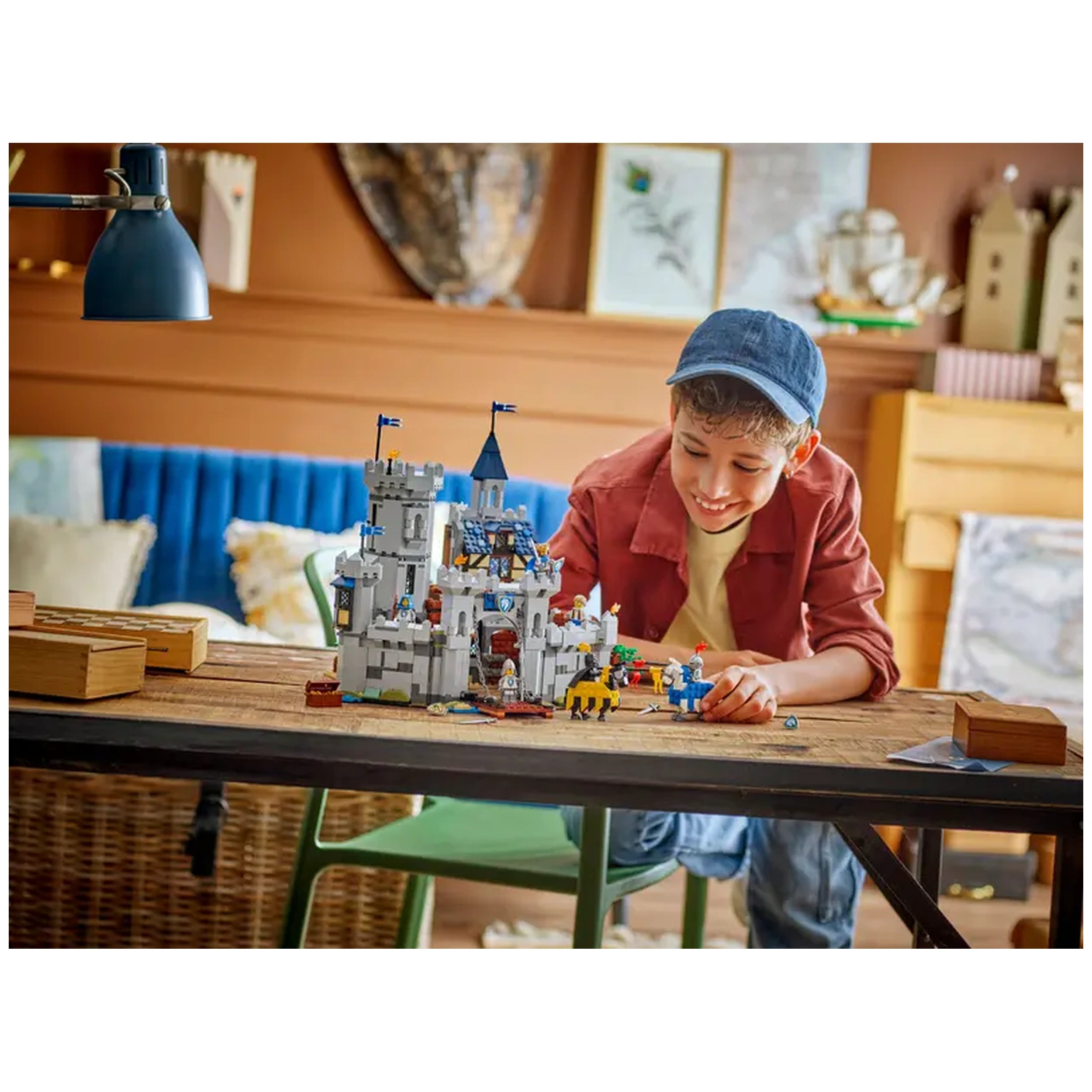 A cheerful boy wearing a blue cap and a reddish jacket sits at a wooden table, intently examining the LEGO Creator 3in1 Medieval Horse Knight Castle playset, which features a detailed castle with towers, flags, and figures. The background includes cozy decor, enhancing the imaginative play atmosphere.