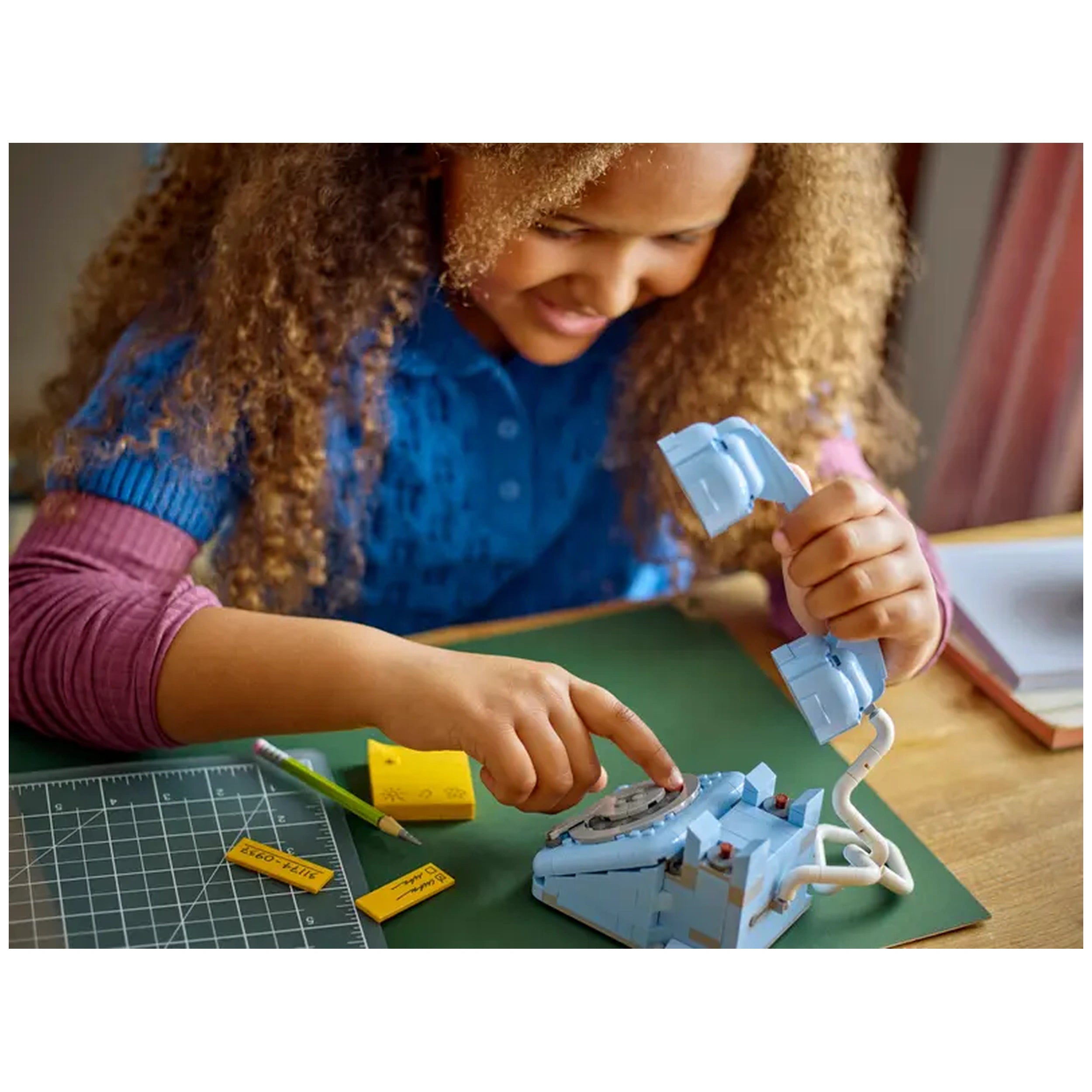 A young girl with curly hair interacts with a blue LEGO Creator 3in1 Retro Telephone set on a green mat. She is lifting the receiver with one hand while pointing at the rotary dial with the other. Nearby, a yellow pencil and sticky notes add color to the workspace.
