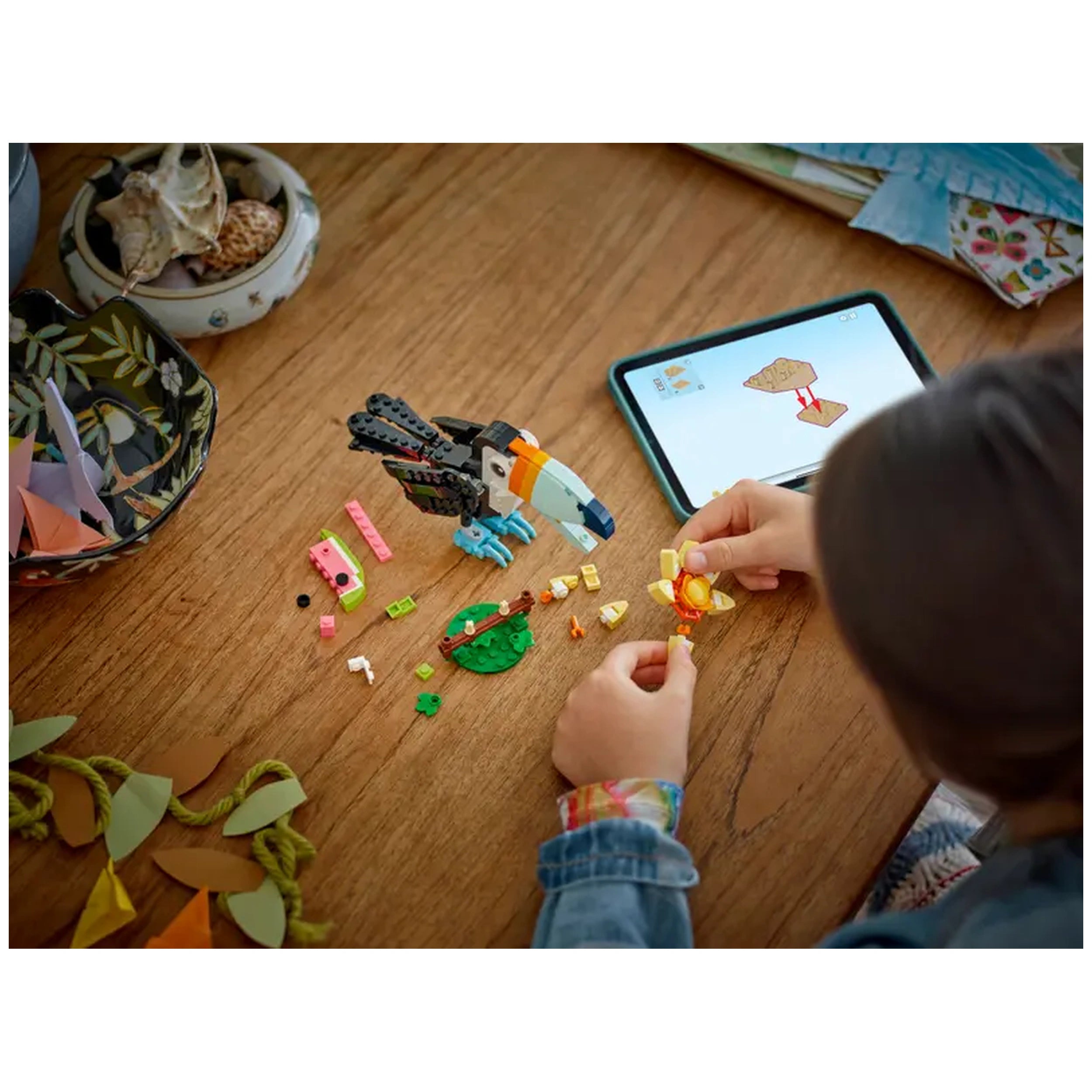 A child assembles colorful LEGO bricks on a wooden table, creating a tropical toucan while using a tablet for guidance.