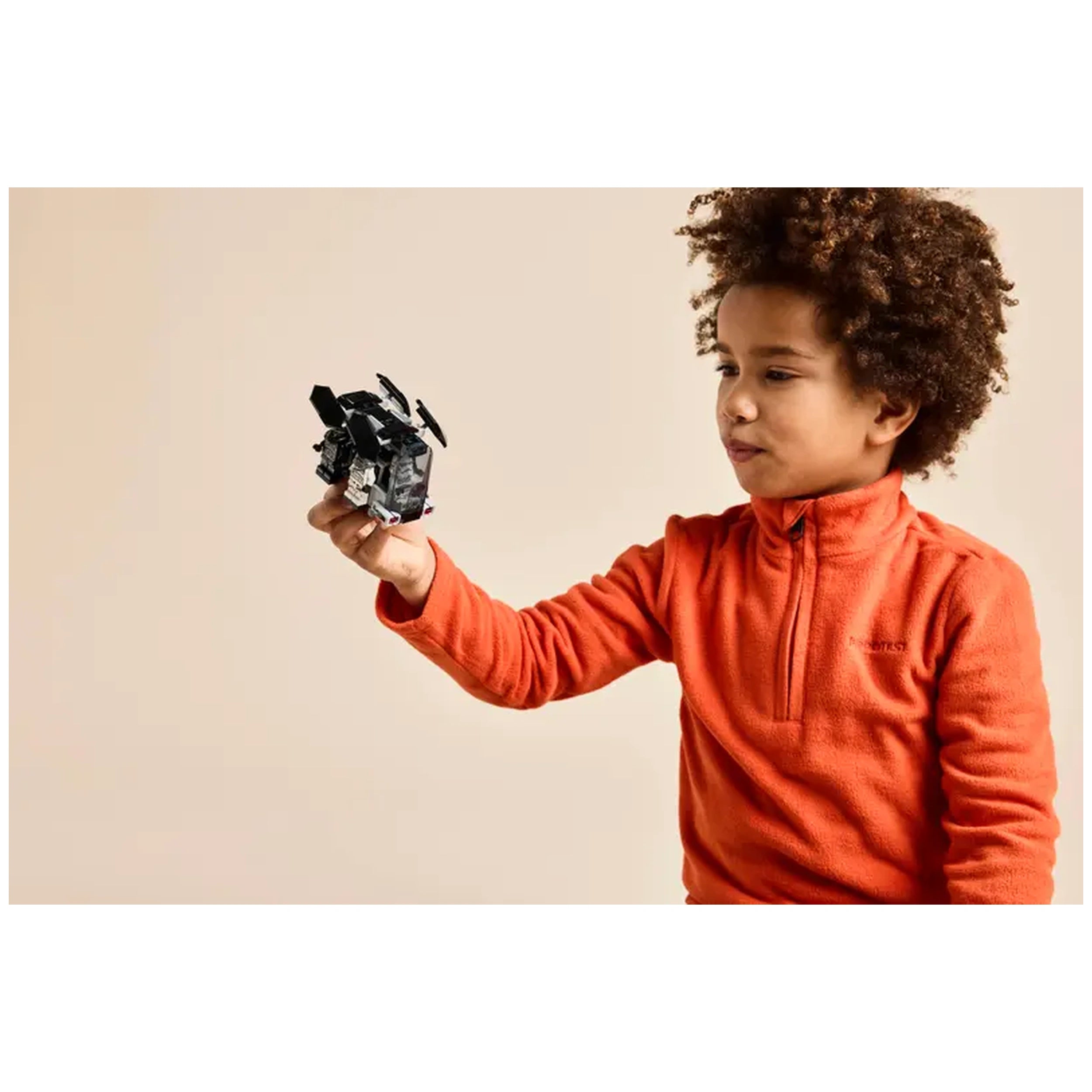 A young boy with curly hair in an orange fleece pullover smiles while holding a LEGO Death Trooper dropship in a playful pose.