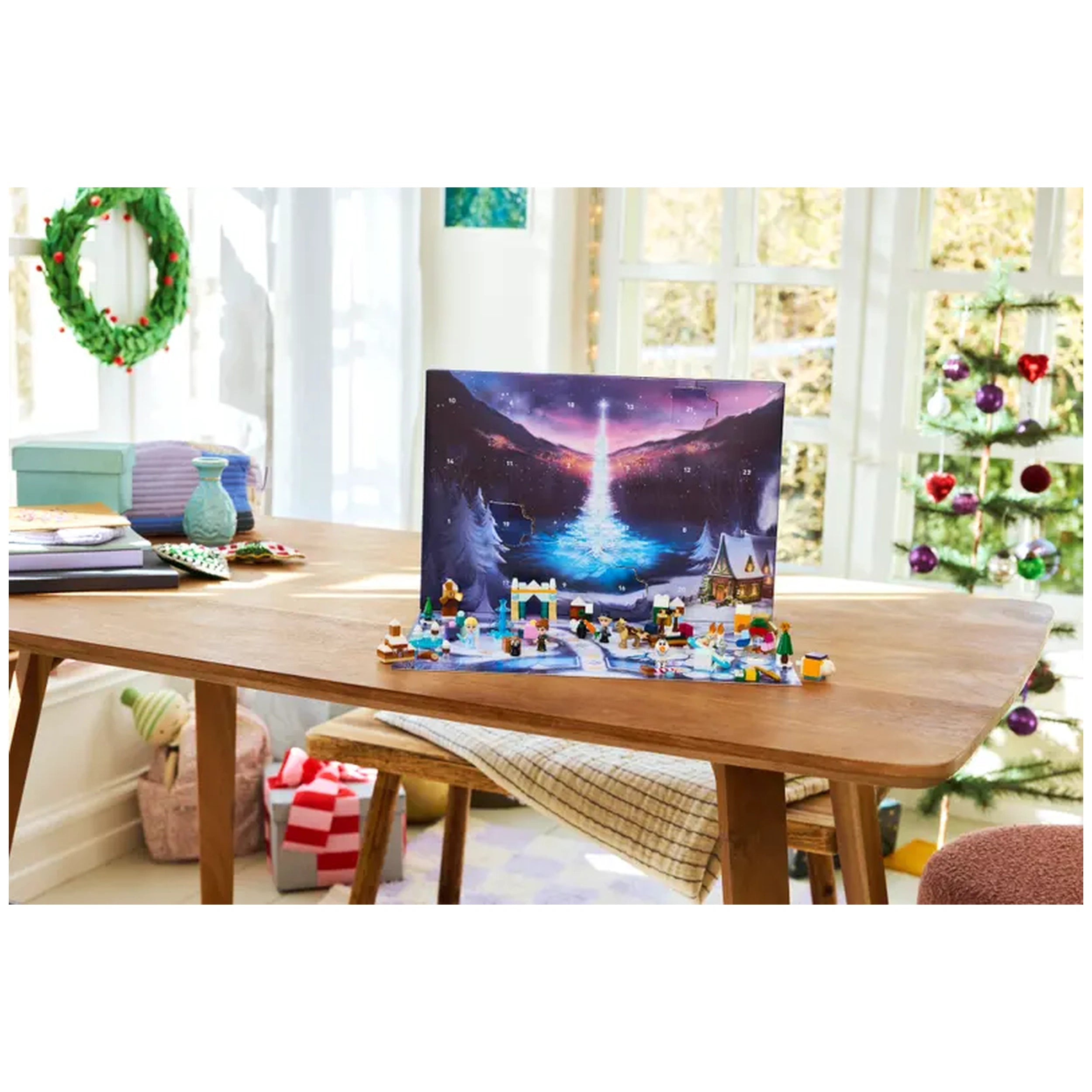 Colorful LEGO Disney Frozen Advent Calendar 2025 displayed on a wooden table, featuring snowy scenery and playful mini figures, with a festive Christmas tree in the background.