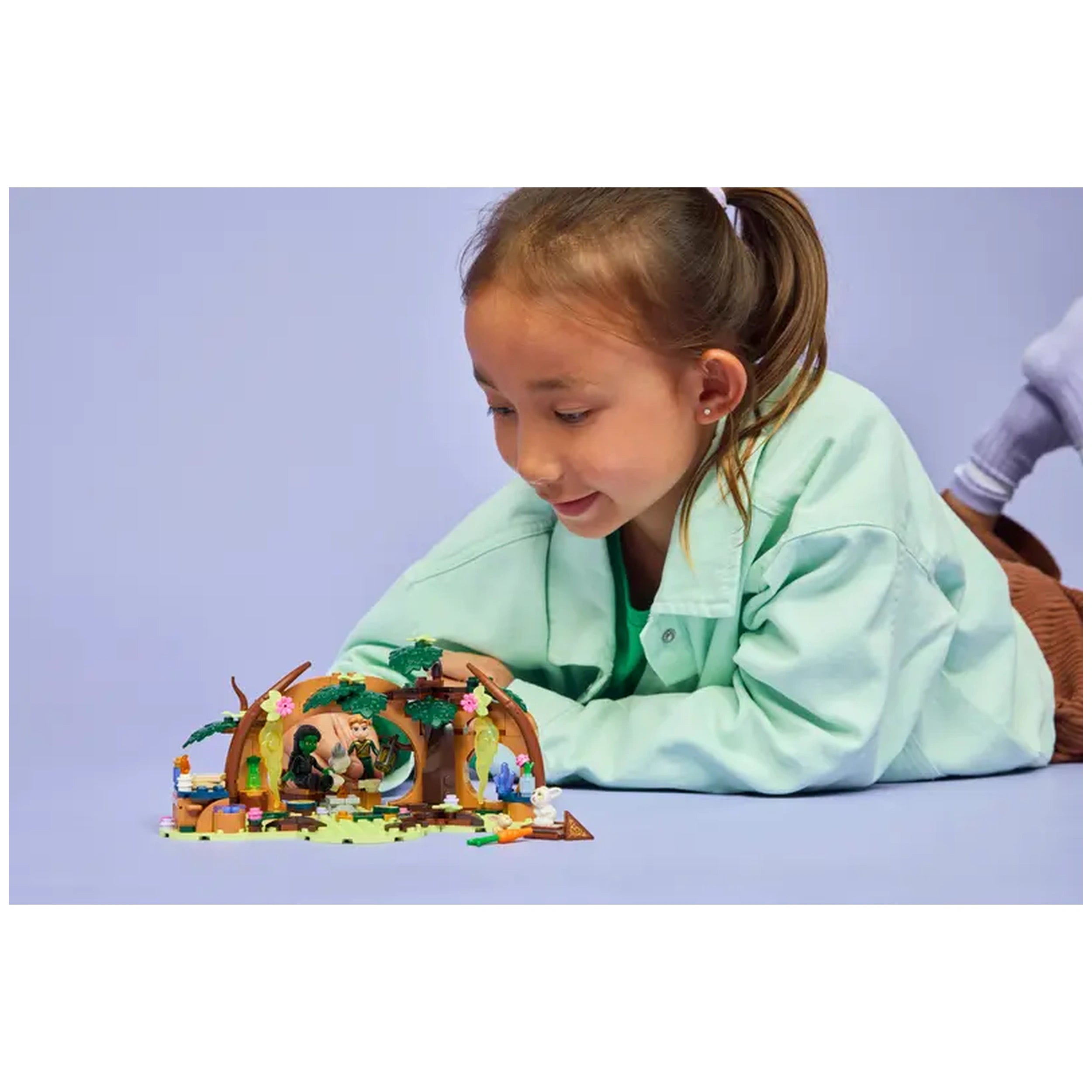 A child in a mint green shirt smiles while playing with the LEGO Elphaba's Retreat set, featuring whimsical trees, colorful details, and characters from the Wicked musical.