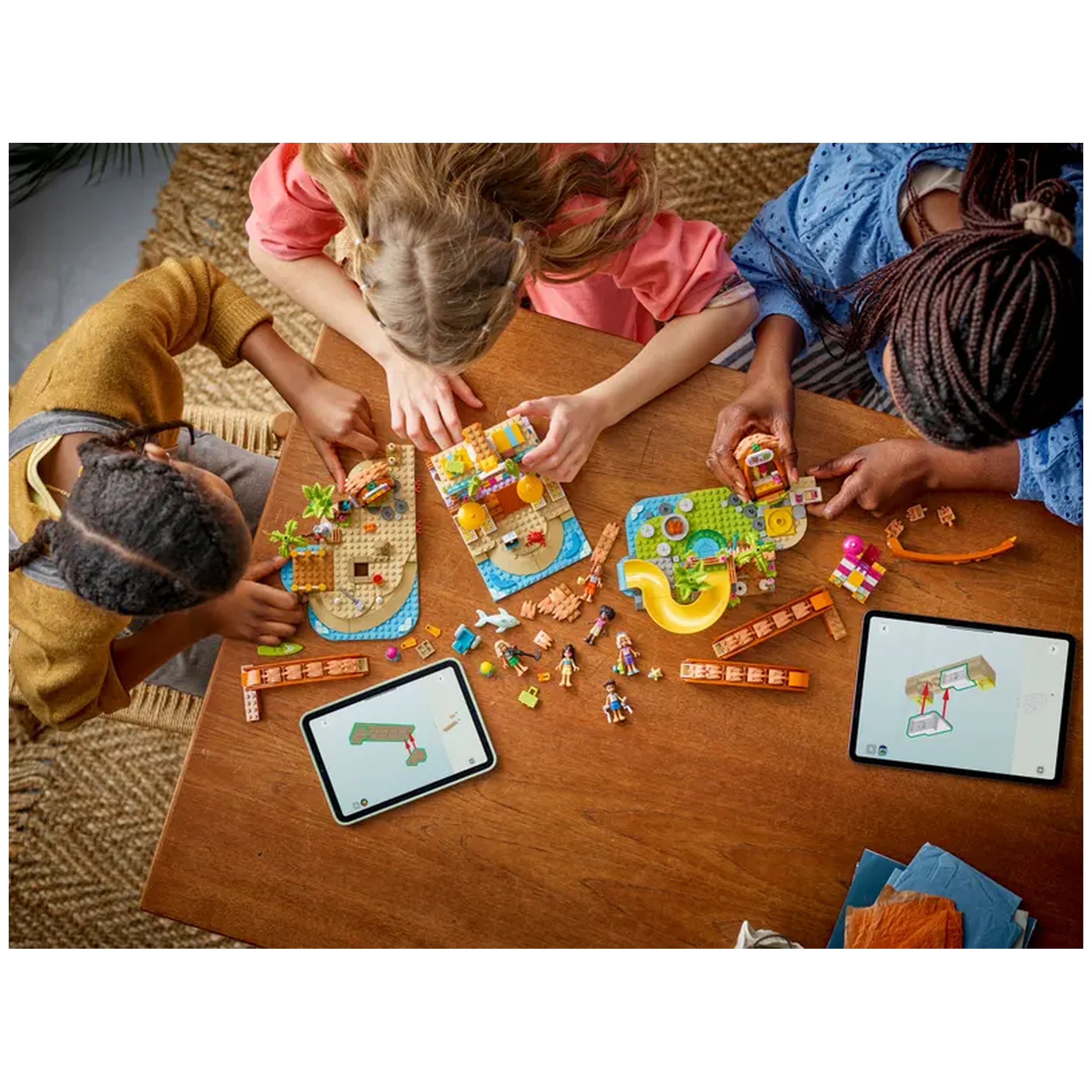Four children collaboratively building a LEGO beach resort set on a wooden table, surrounded by colorful pieces and two tablets displaying digital instructions.